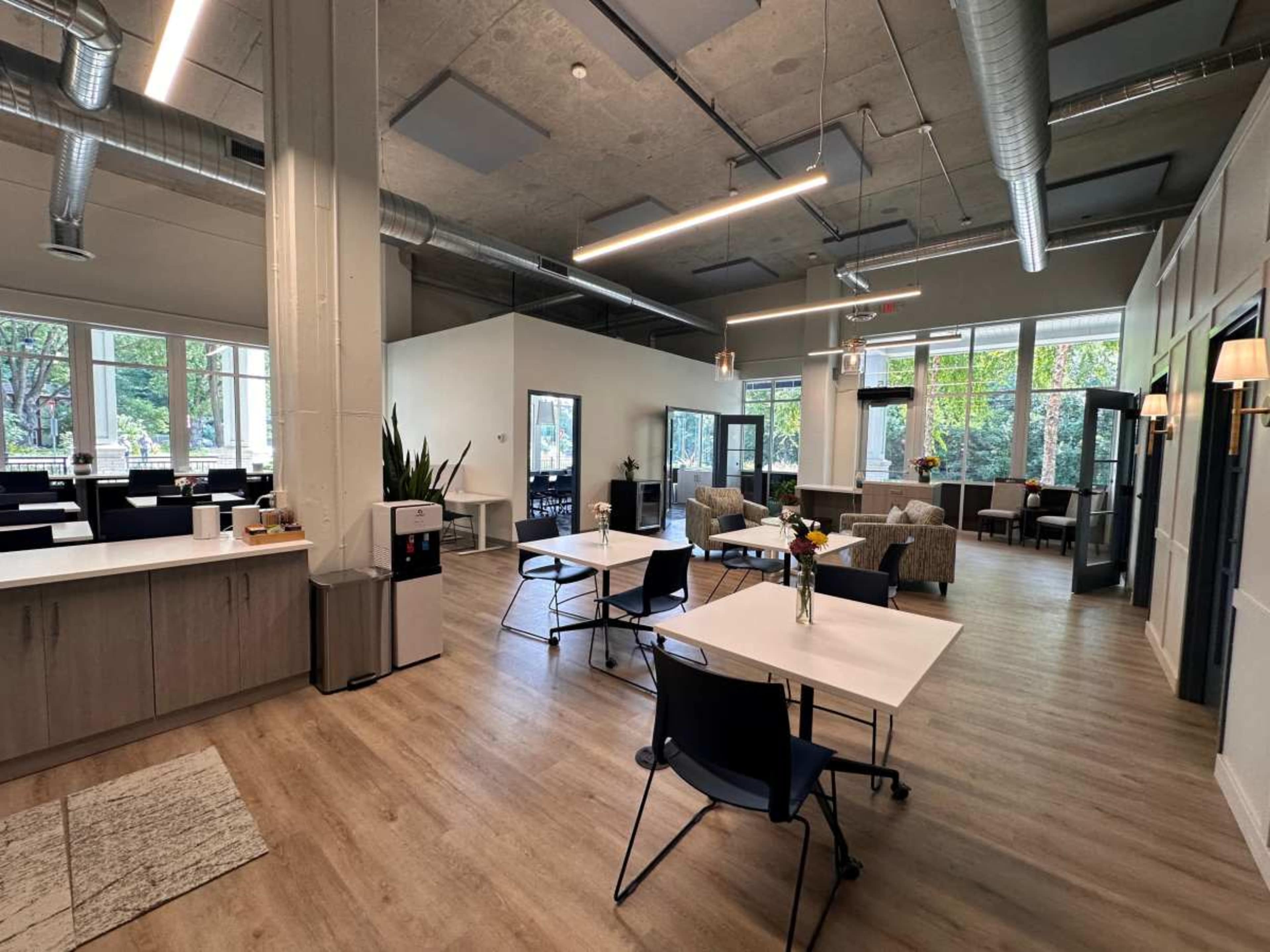 The image shows a modern open-space office or communal area featuring tables, chairs, and a seating arrangement with large windows allowing natural light.