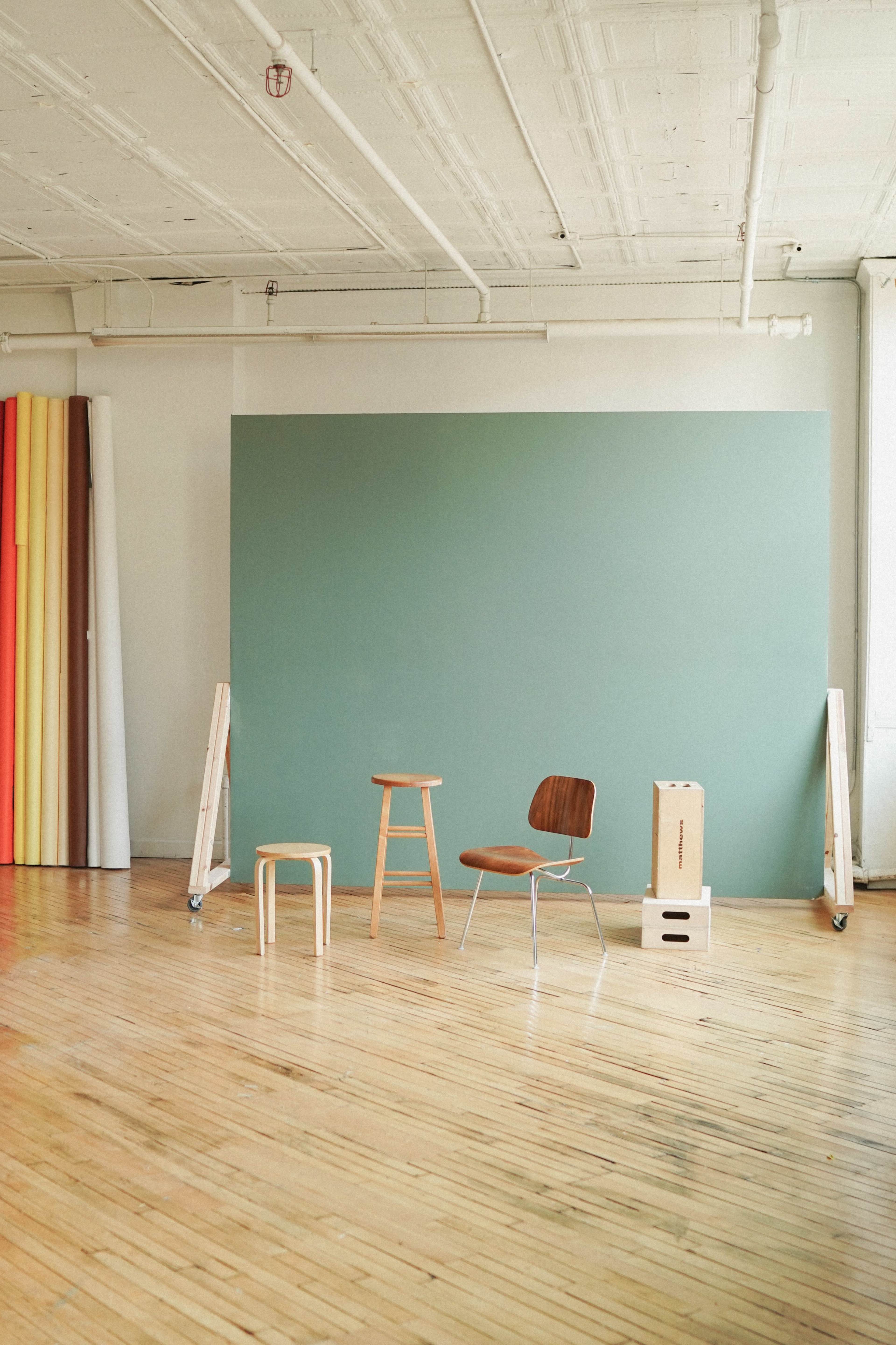 The scene features a minimalist studio space with a green backdrop, three wooden stools or chairs, and colorful rolls of backdrop materials leaning against the wall.