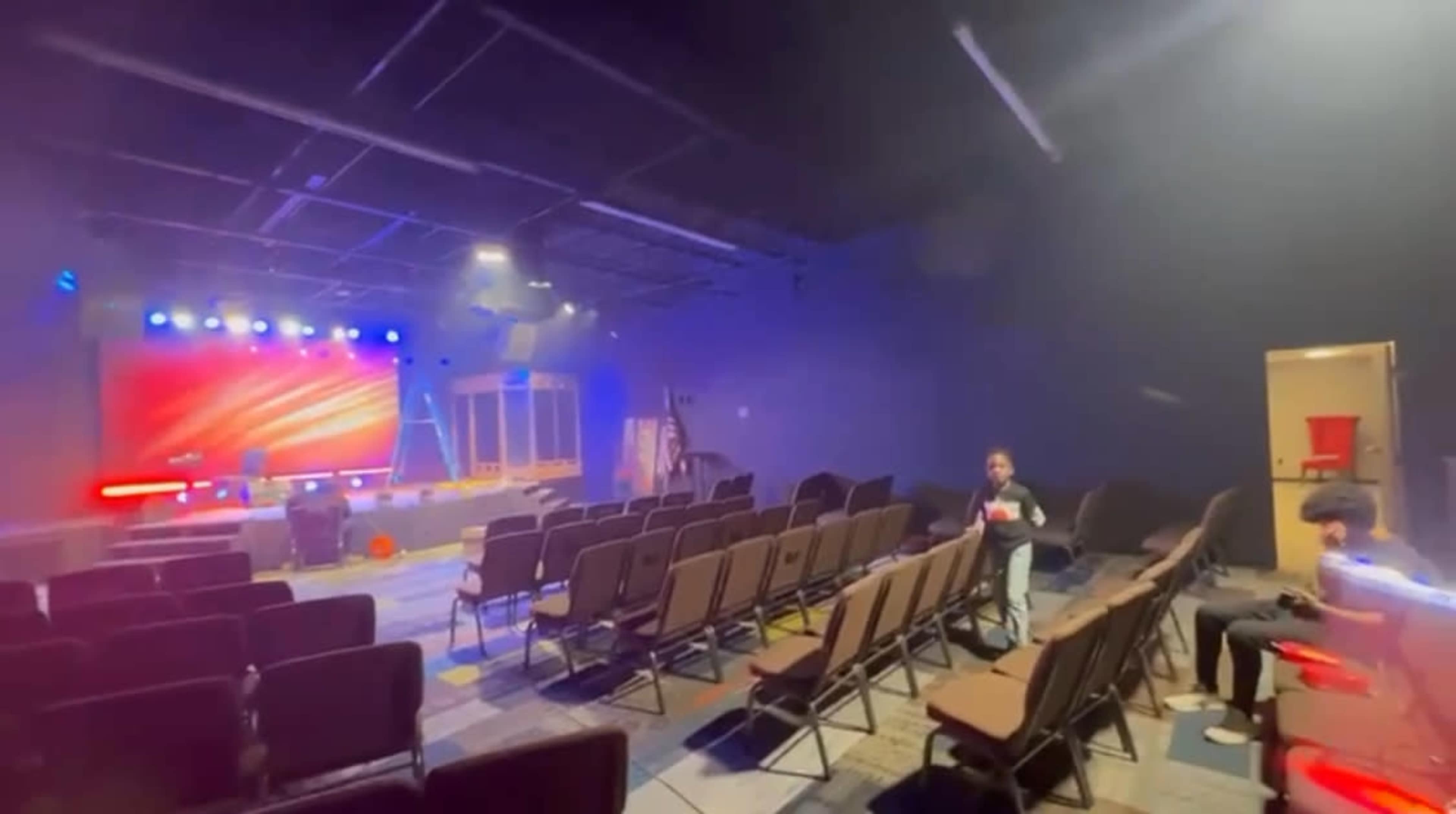 A dimly lit auditorium with rows of empty chairs facing a brightly lit stage, and a few people preparing equipment in the background.