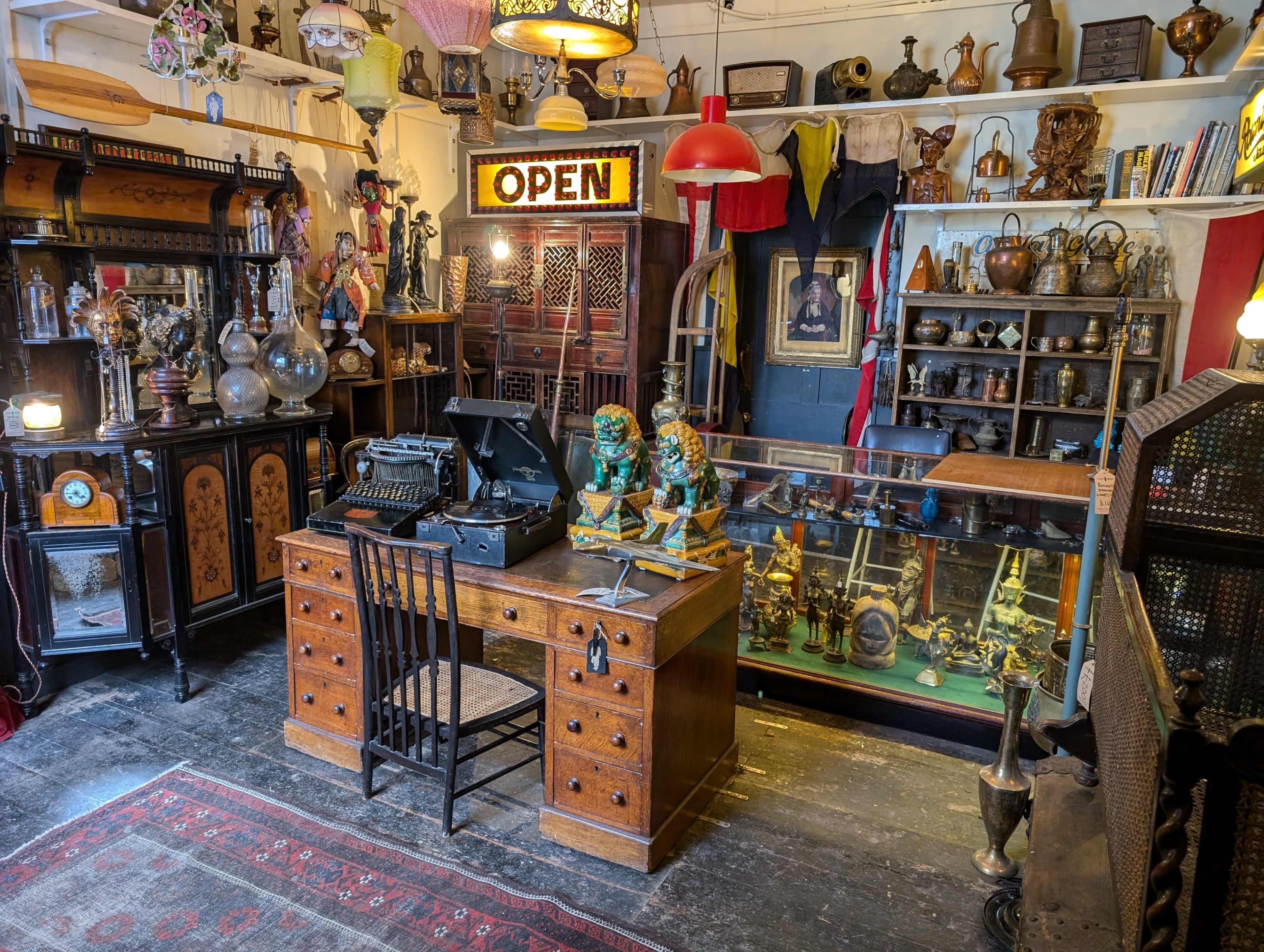 The image shows an antique shop featuring a wooden desk with a typewriter, illuminated by a neon "OPEN" sign and various vintage items displayed throughout the room.