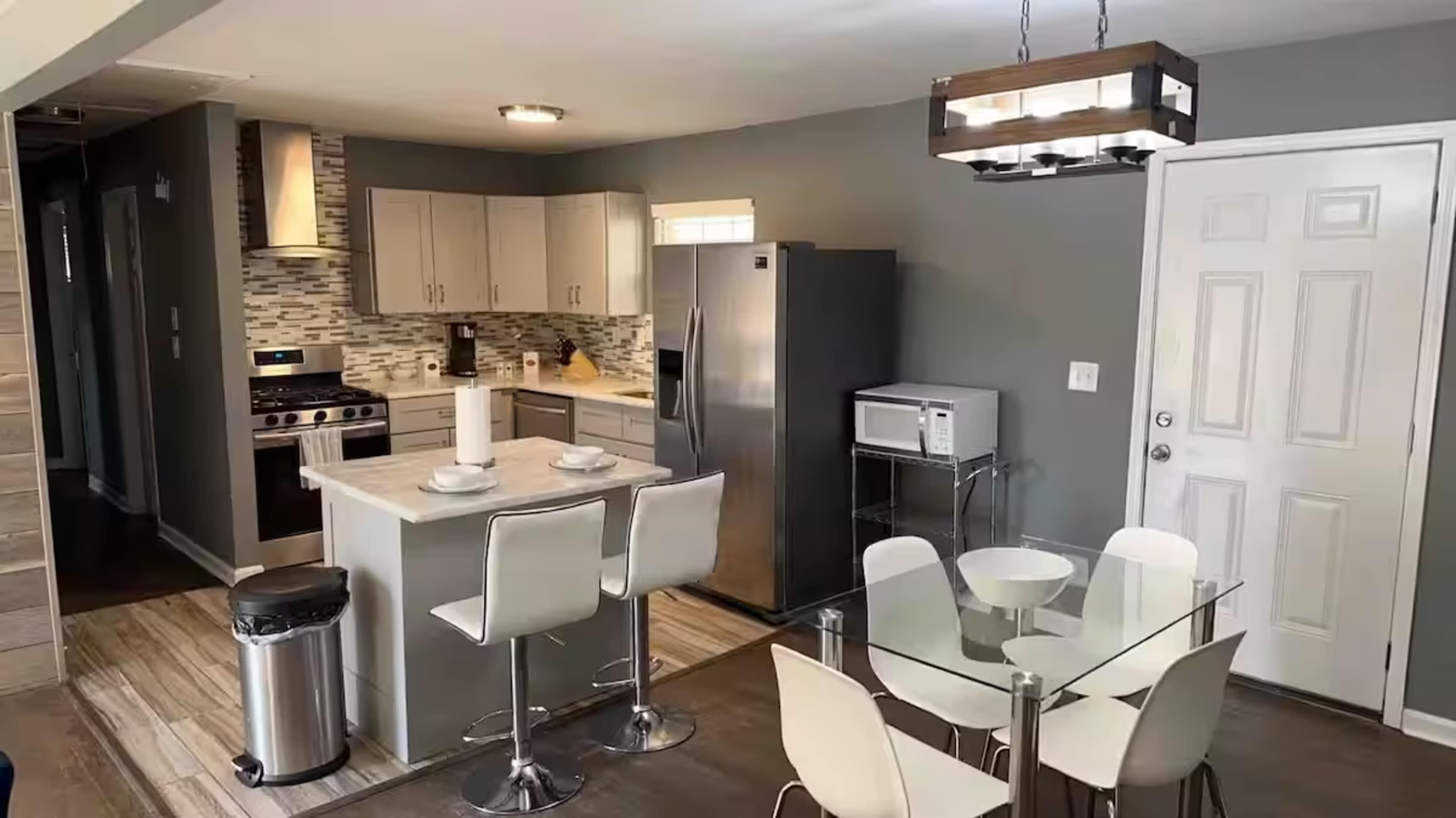 The image shows a modern kitchen and dining area featuring stainless steel appliances, a glass dining table, and light-colored cabinets against a gray wall.