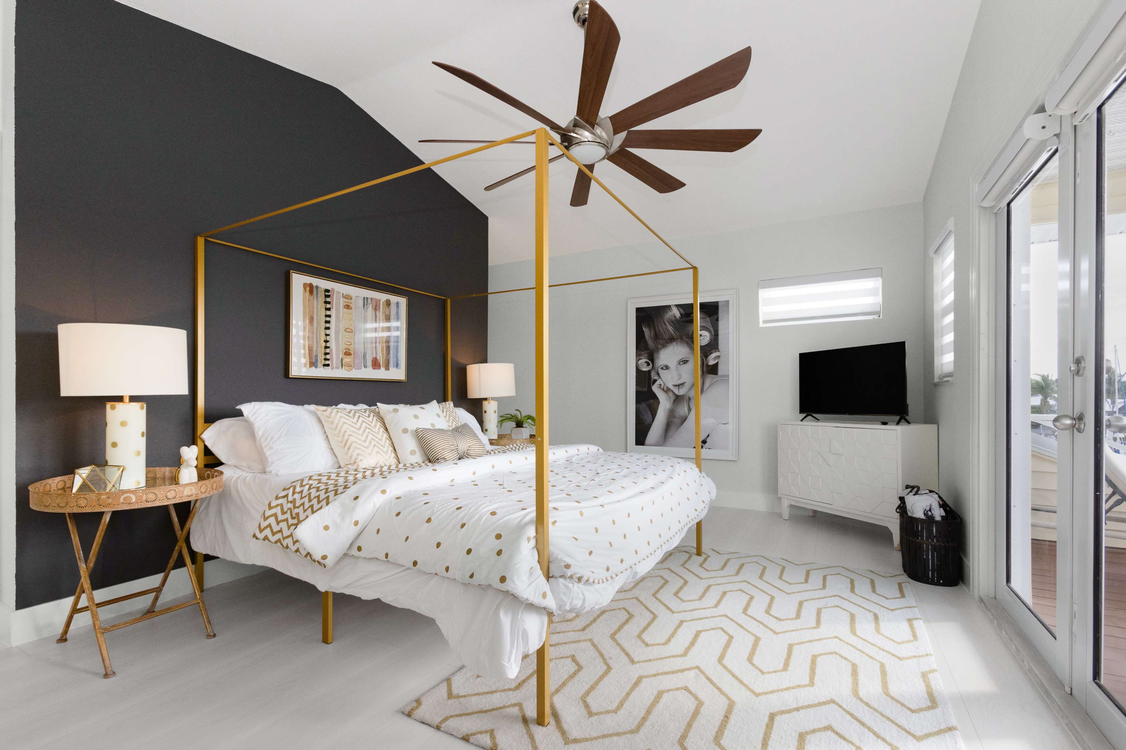 A modern bedroom features a golden four-poster bed, a patterned rug, and a minimalist television stand, complemented by wall art and a ceiling fan.
