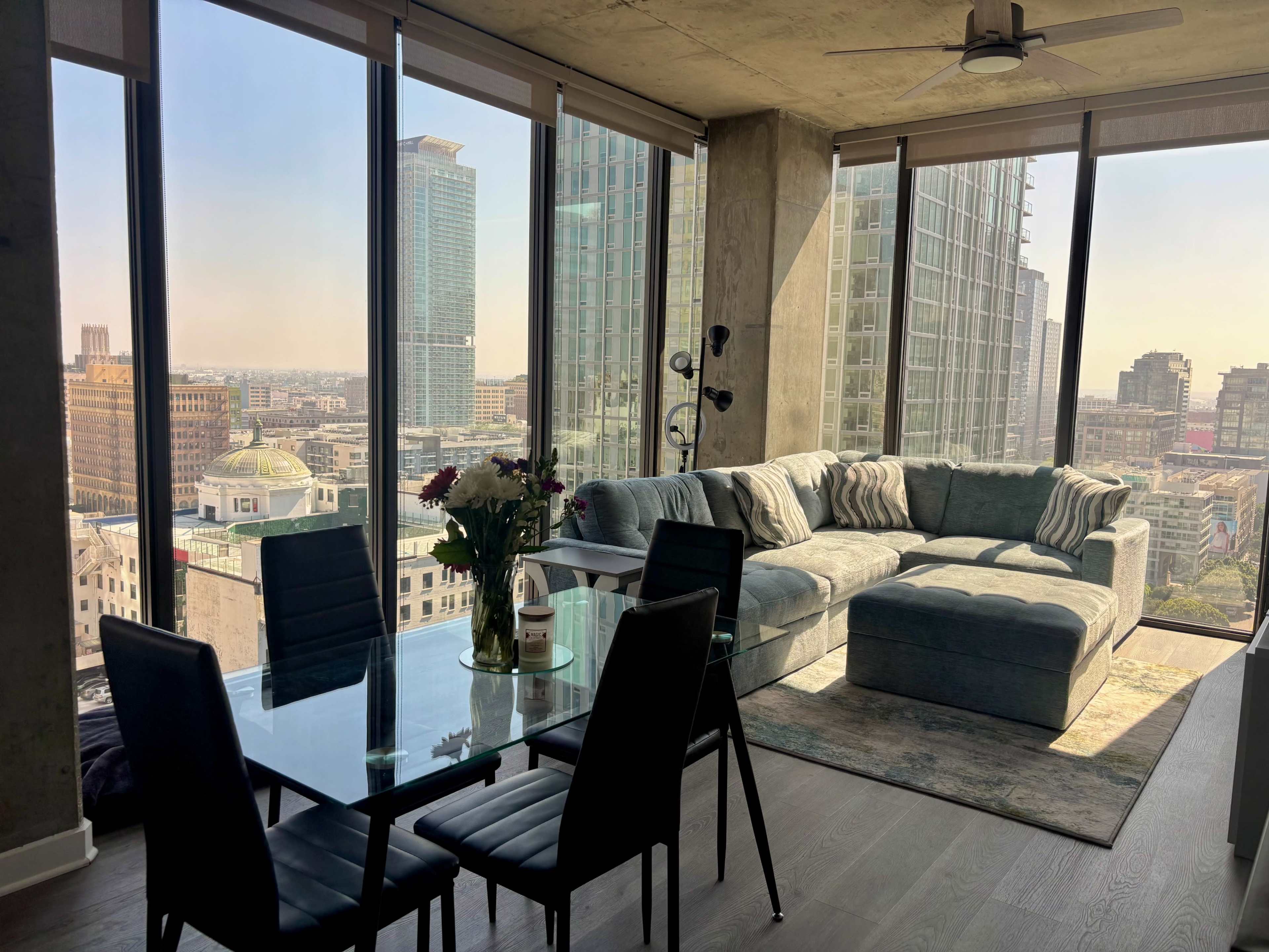 A modern living area features a glass dining table and a sectional sofa, with large windows providing a view of the city skyline.