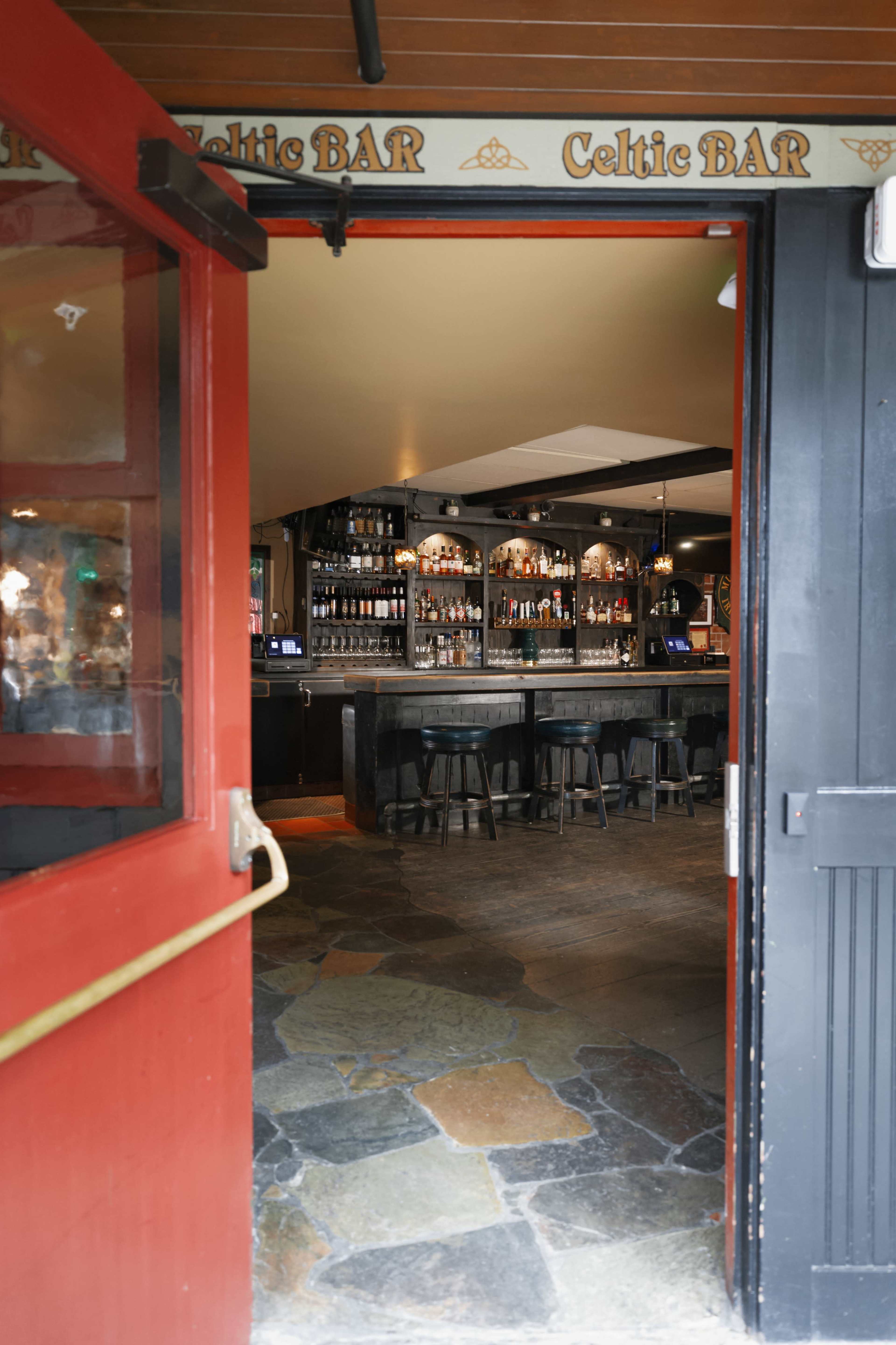 A red door opens to reveal a dimly lit bar with wooden shelves and a dark countertop lined with stools.