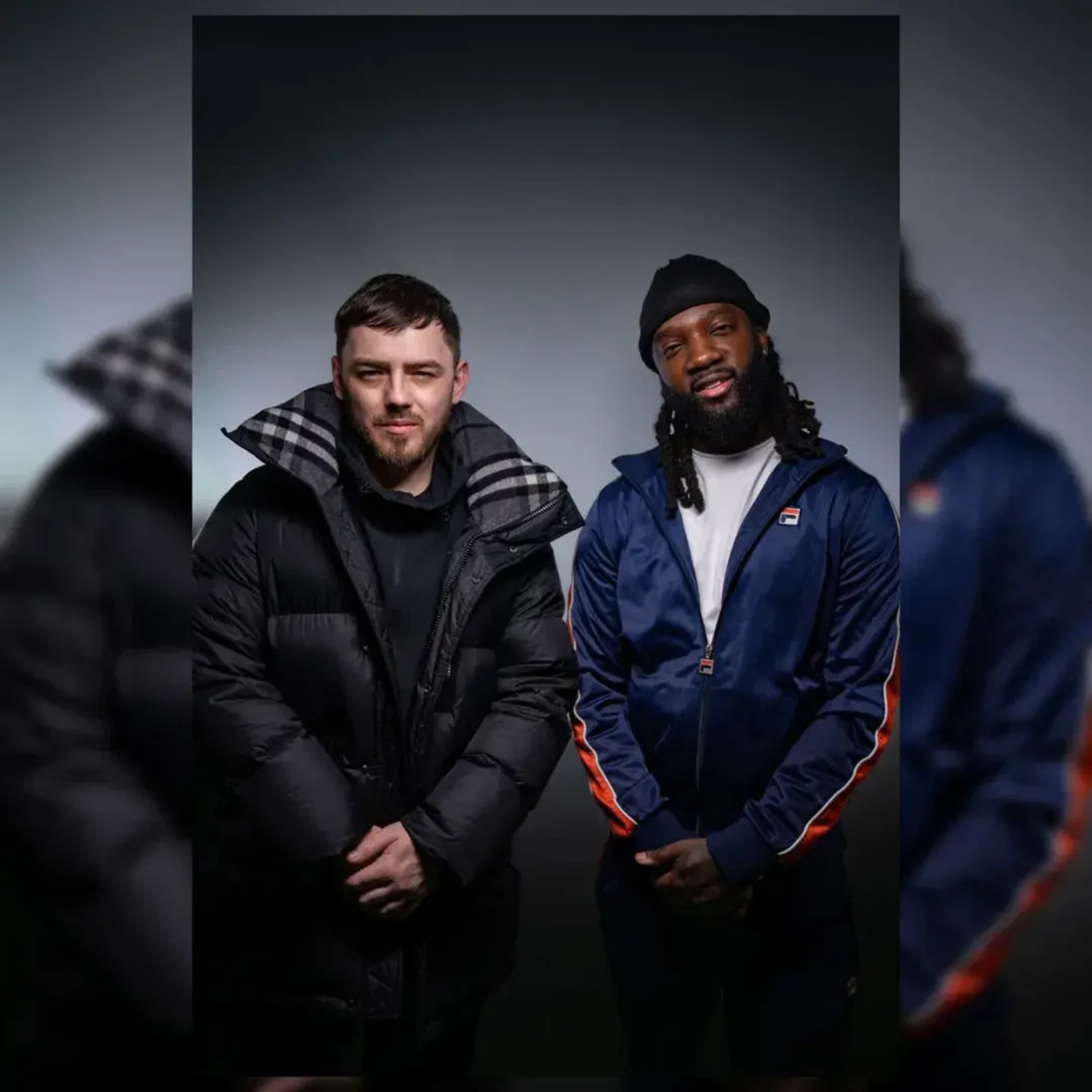 Two men are standing side by side, one in a black puffer jacket and the other in a blue tracksuit with orange accents, posed against a gray background.