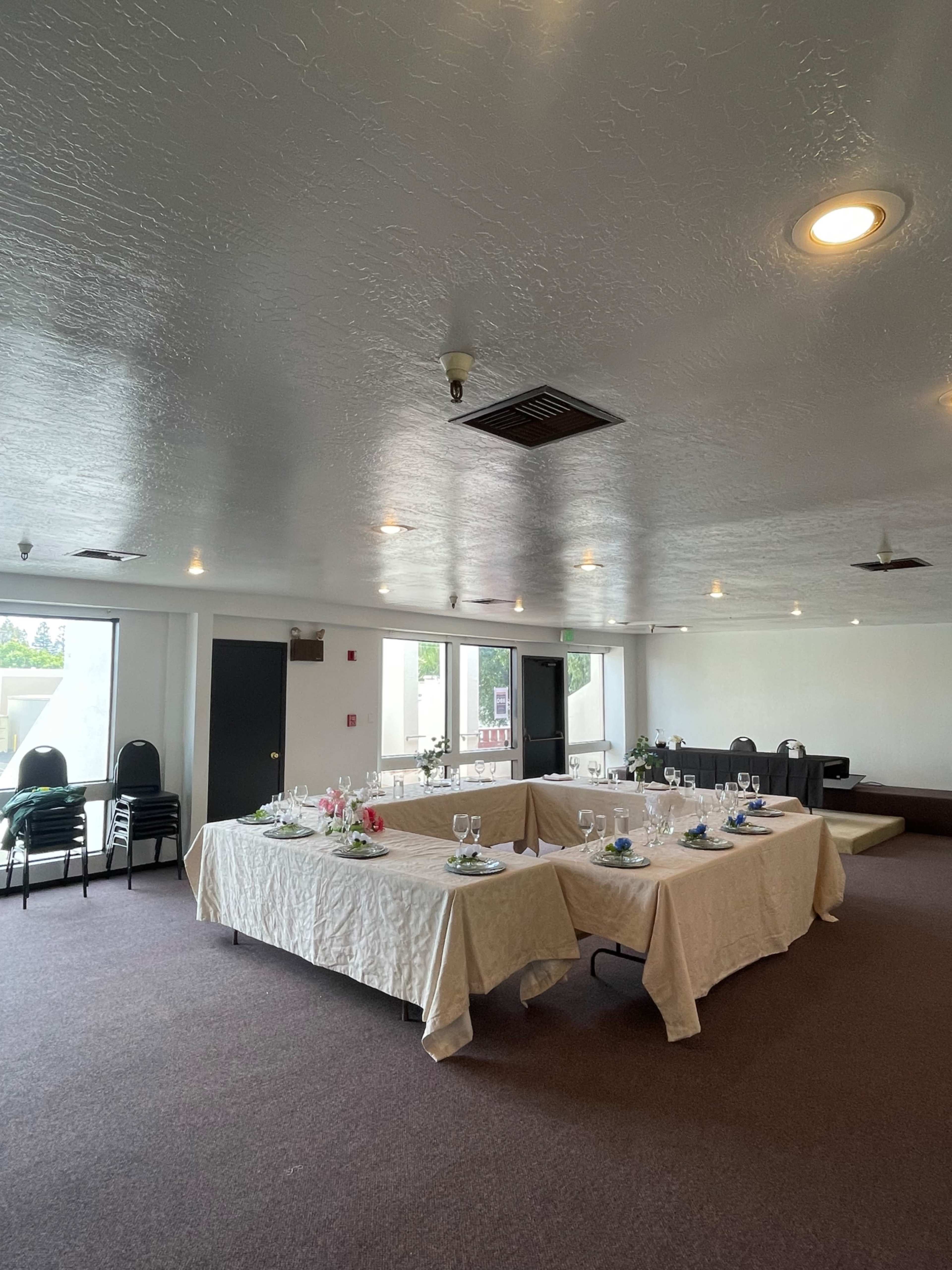 A spacious indoor room arranged with tables set for a meal, featuring white tablecloths and decorative place settings, surrounded by chairs.