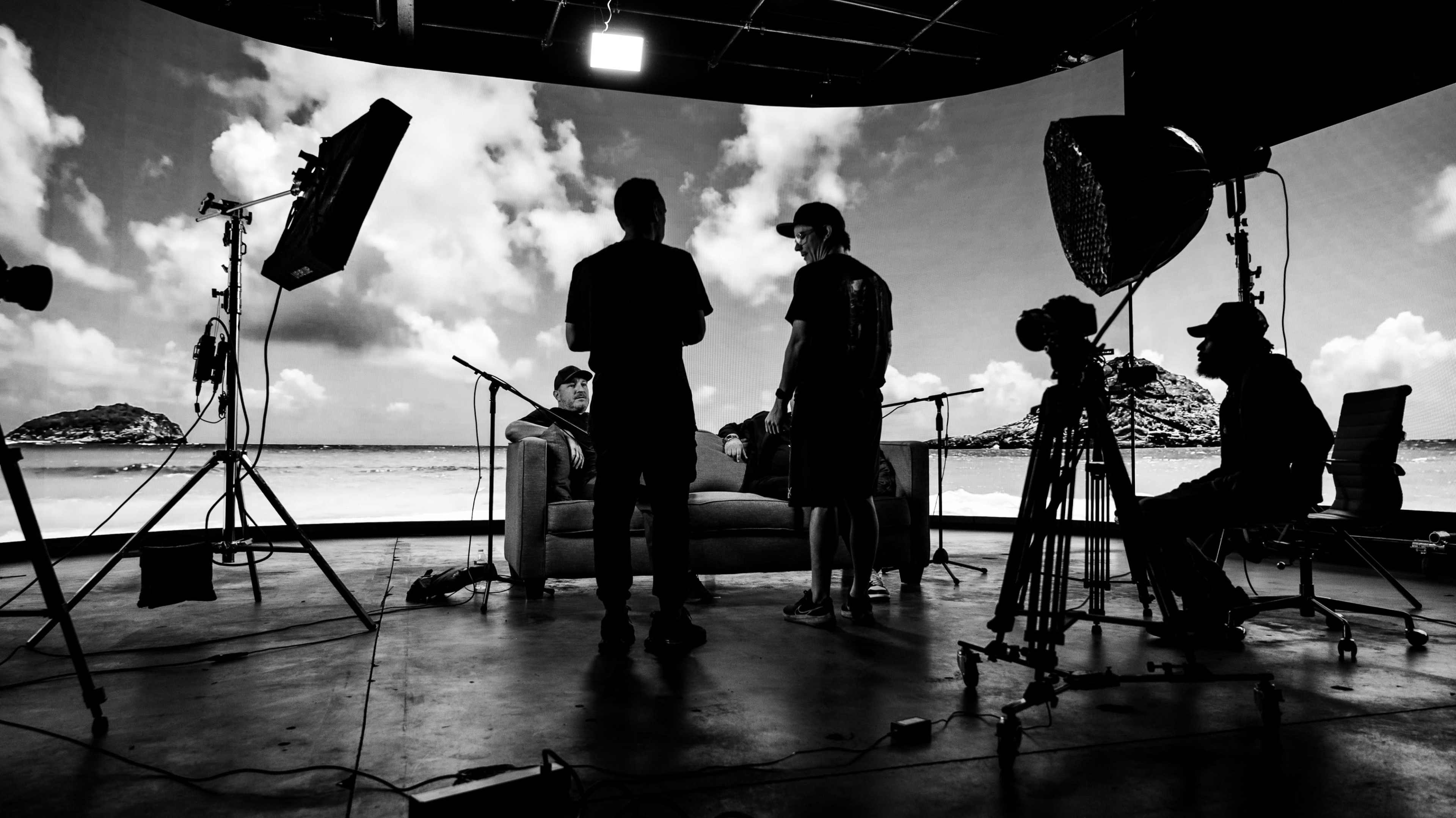A film production crew gathers around a sofa on a set with a large backdrop displaying clouds and a beach scene.
