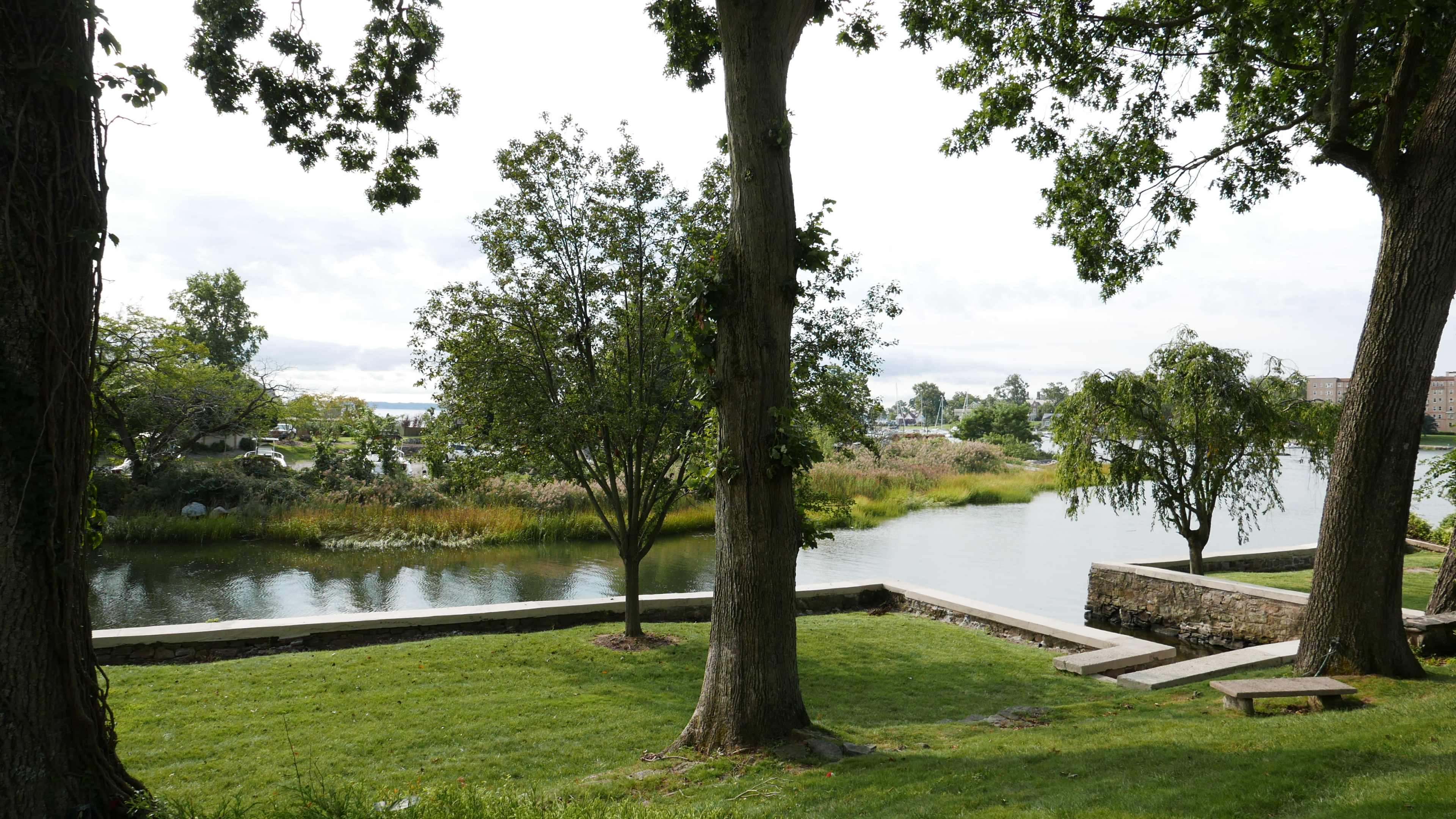 The image shows a tranquil landscape featuring a river bordered by trees and grassy areas, with stone pathways along the shore.