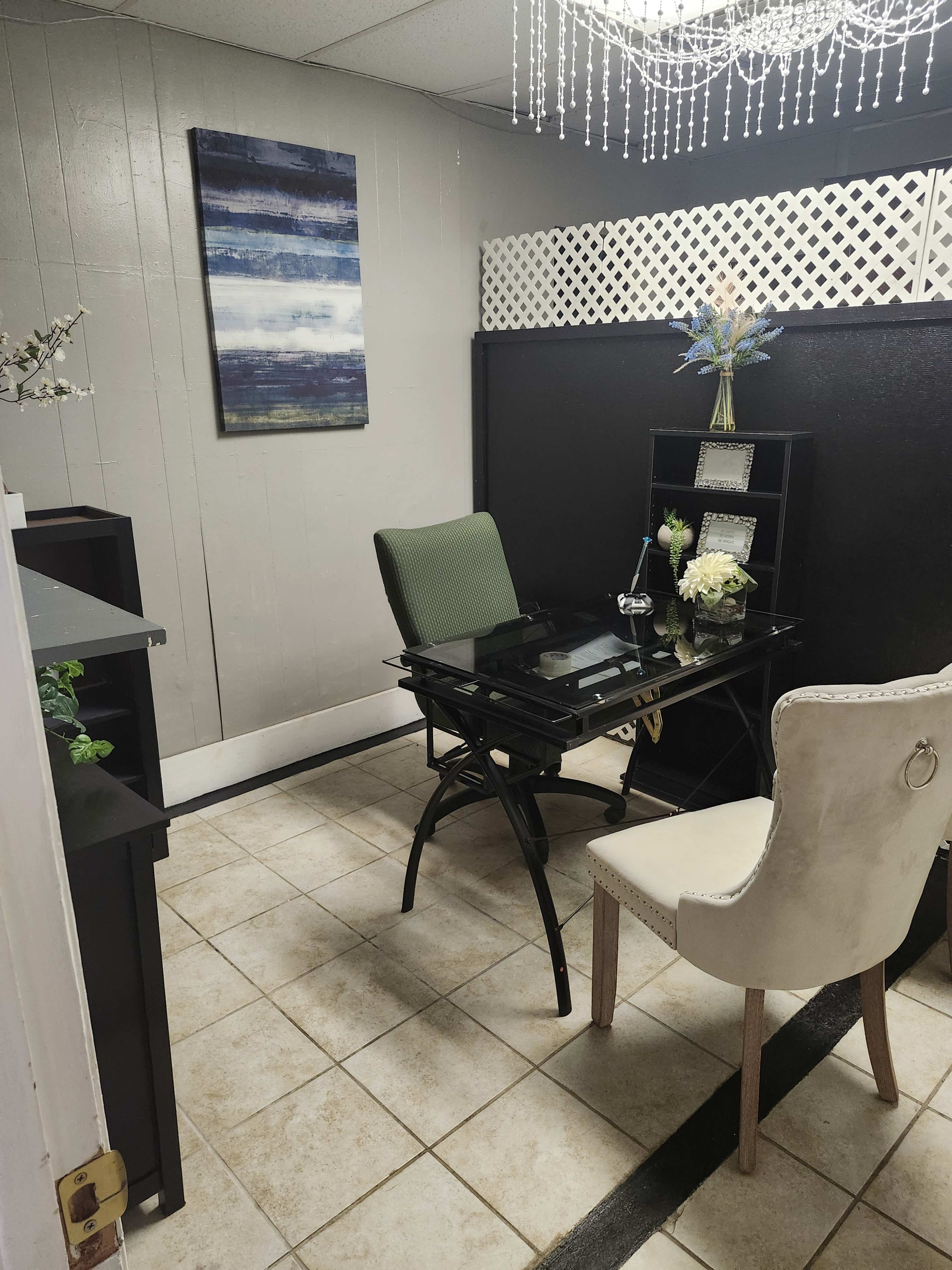 The image shows a small office space with a black desk, a green chair, and a white chair, surrounded by tiled flooring and a decorative wall.