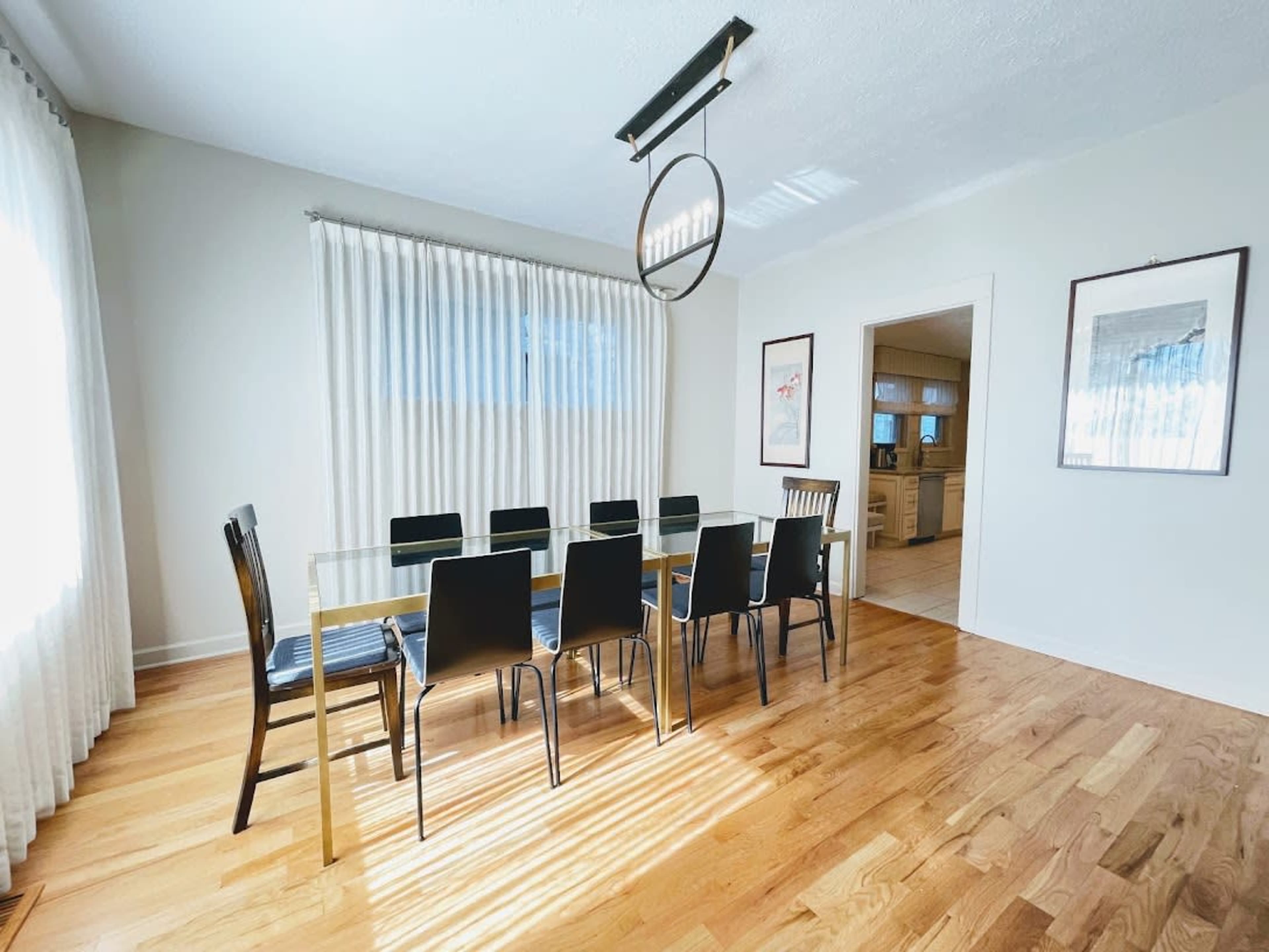 A modern dining room features a long glass table surrounded by black chairs, with natural light streaming through sheer curtains.