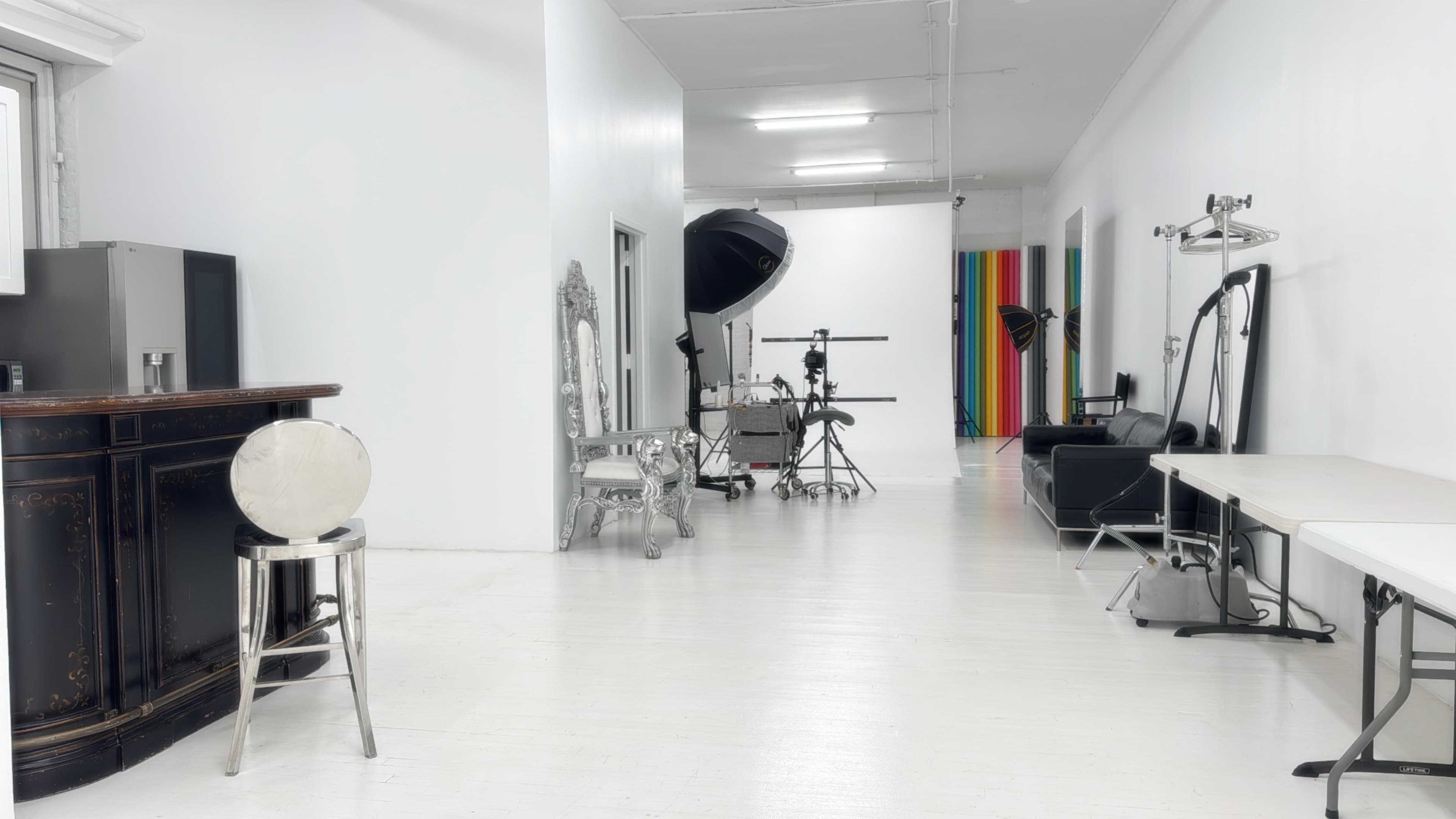 The image shows a spacious, empty studio with white walls, a bright floor, and various photography equipment arranged along the sides.