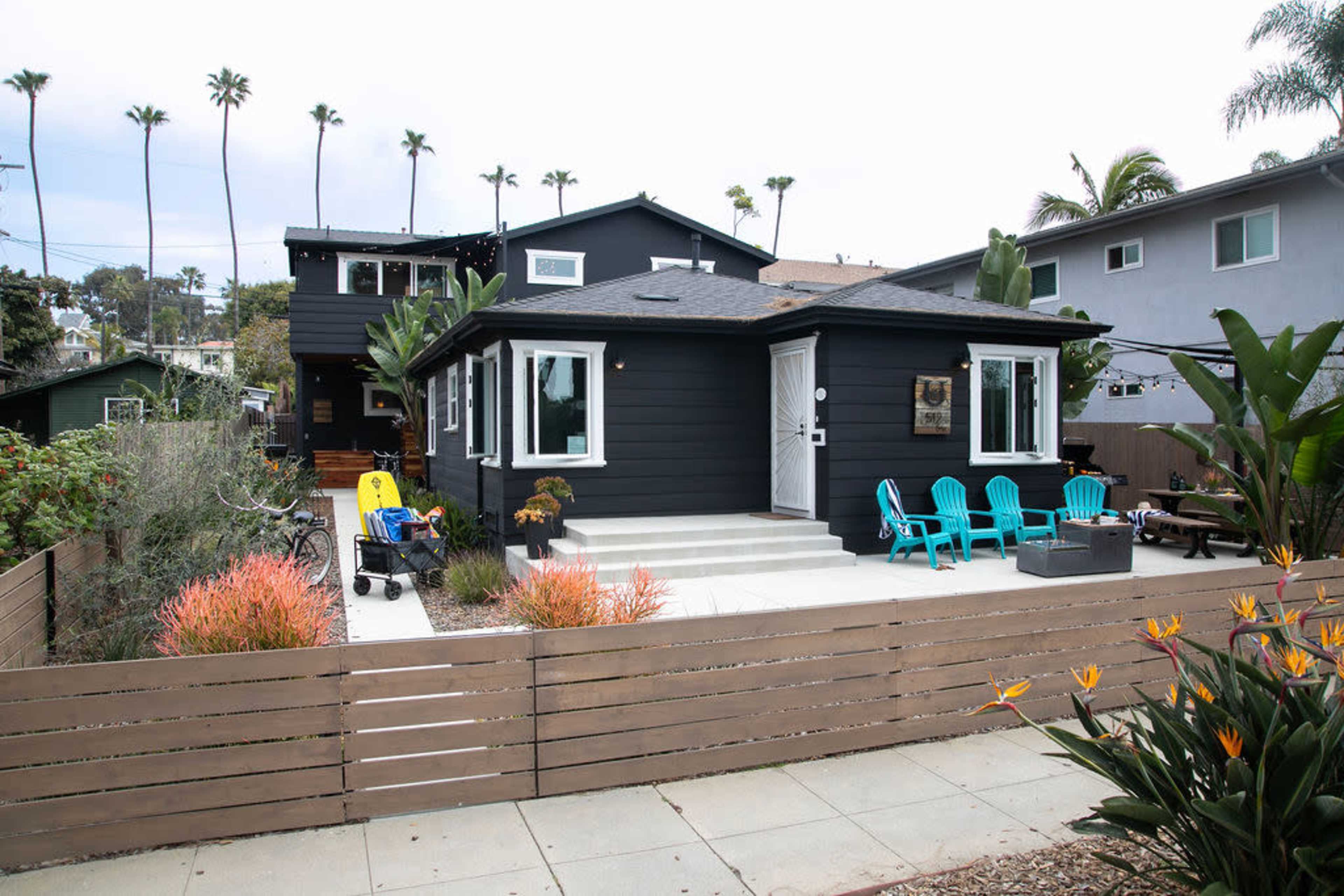 The image shows a modern black house with a white porch and turquoise chairs, surrounded by landscaped gardens and palm trees in the background.
