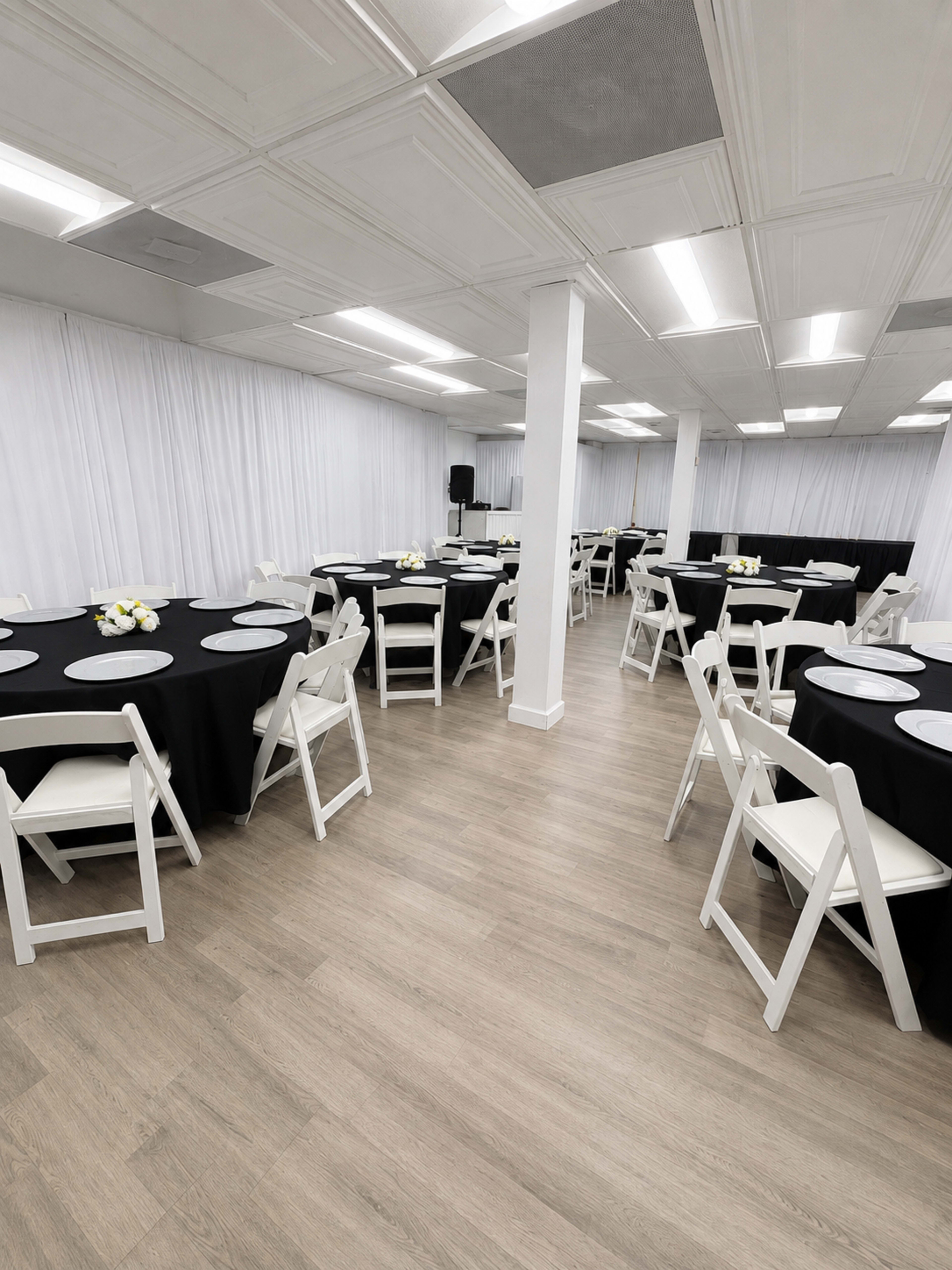The image shows a banquet hall with multiple tables set up, each covered with black tablecloths and decorated with centerpieces, surrounded by white chairs.