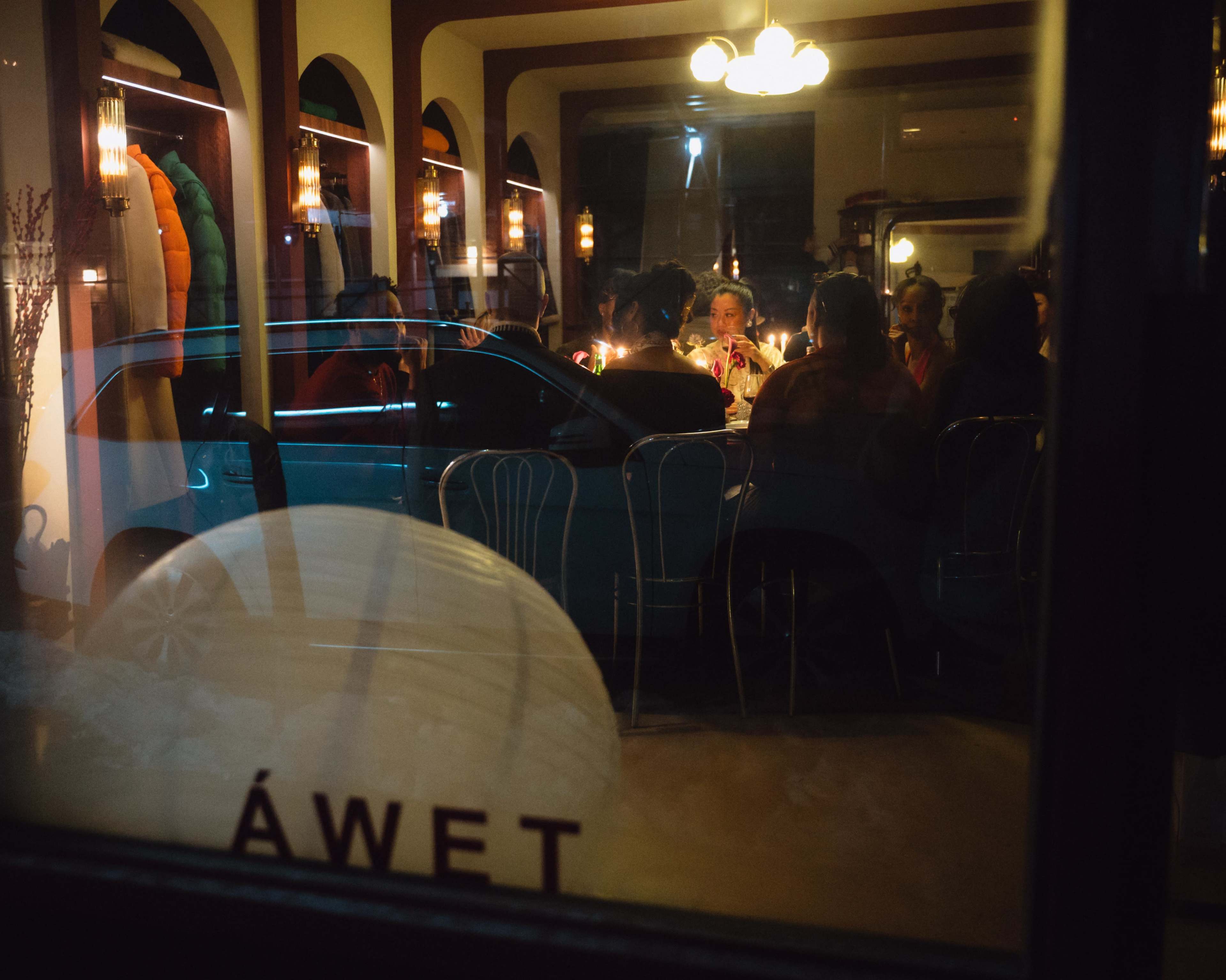 A group of people is seated around a table with candles inside a restaurant, while a car is parked just outside the window.