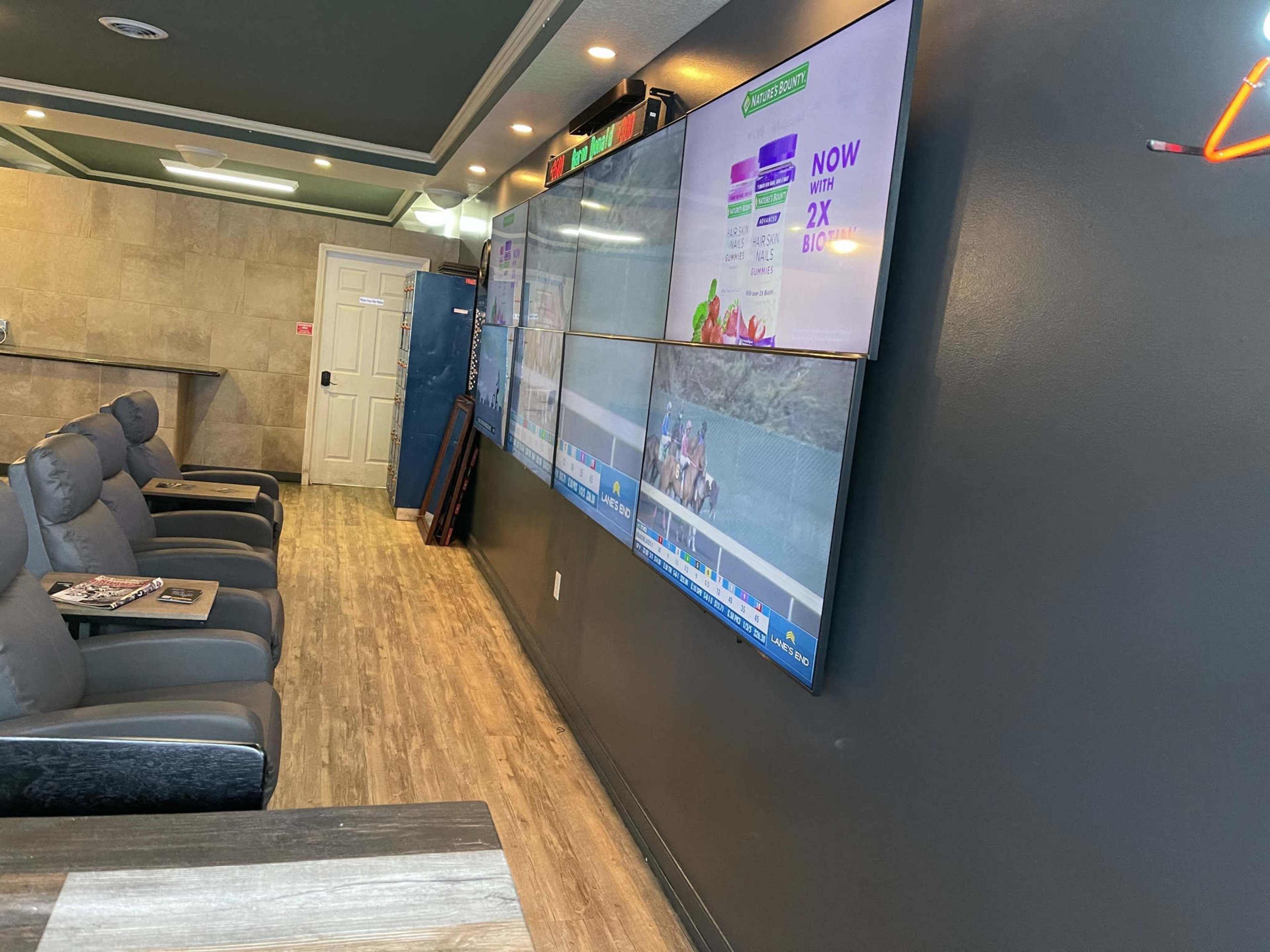 A room with multiple large screens displaying content, facing a row of gray reclining chairs and a brown table.