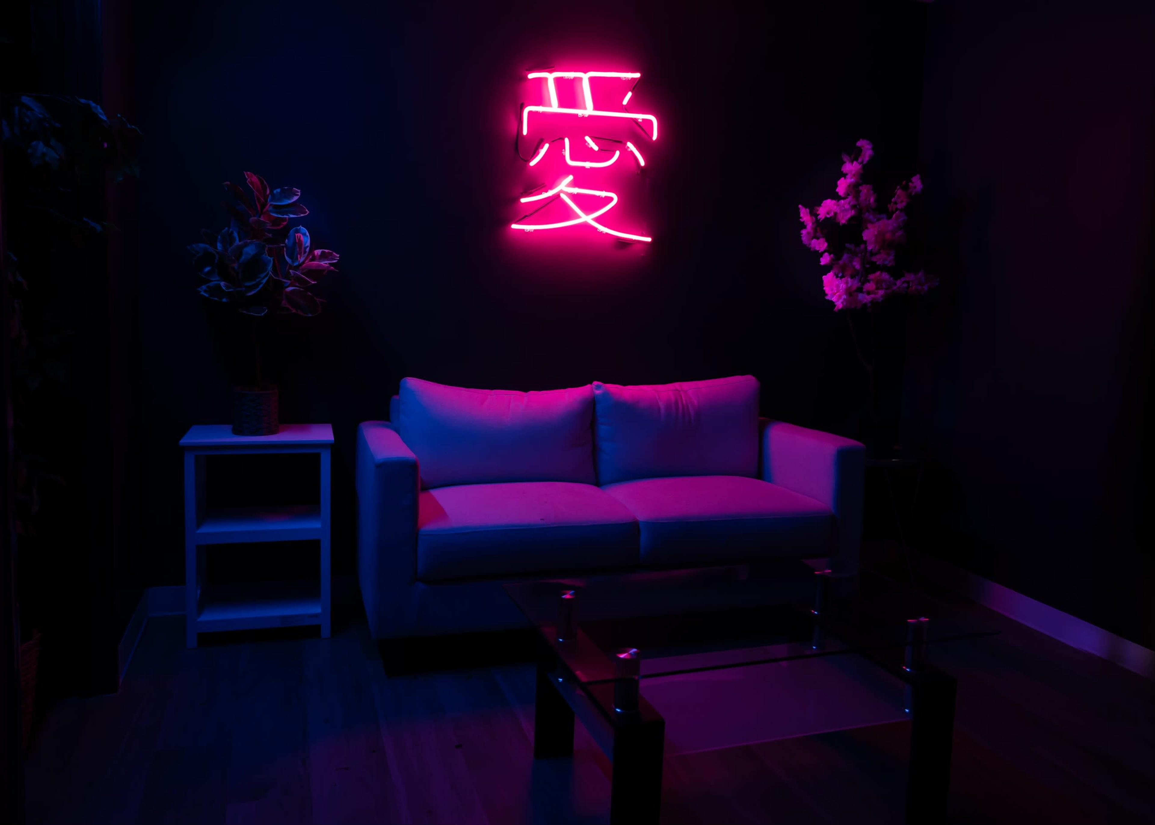 A minimalist lounge area features a light-colored couch, a glass coffee table, and a neon sign with a Chinese character, all set against a dark wall adorned with decorative plants.