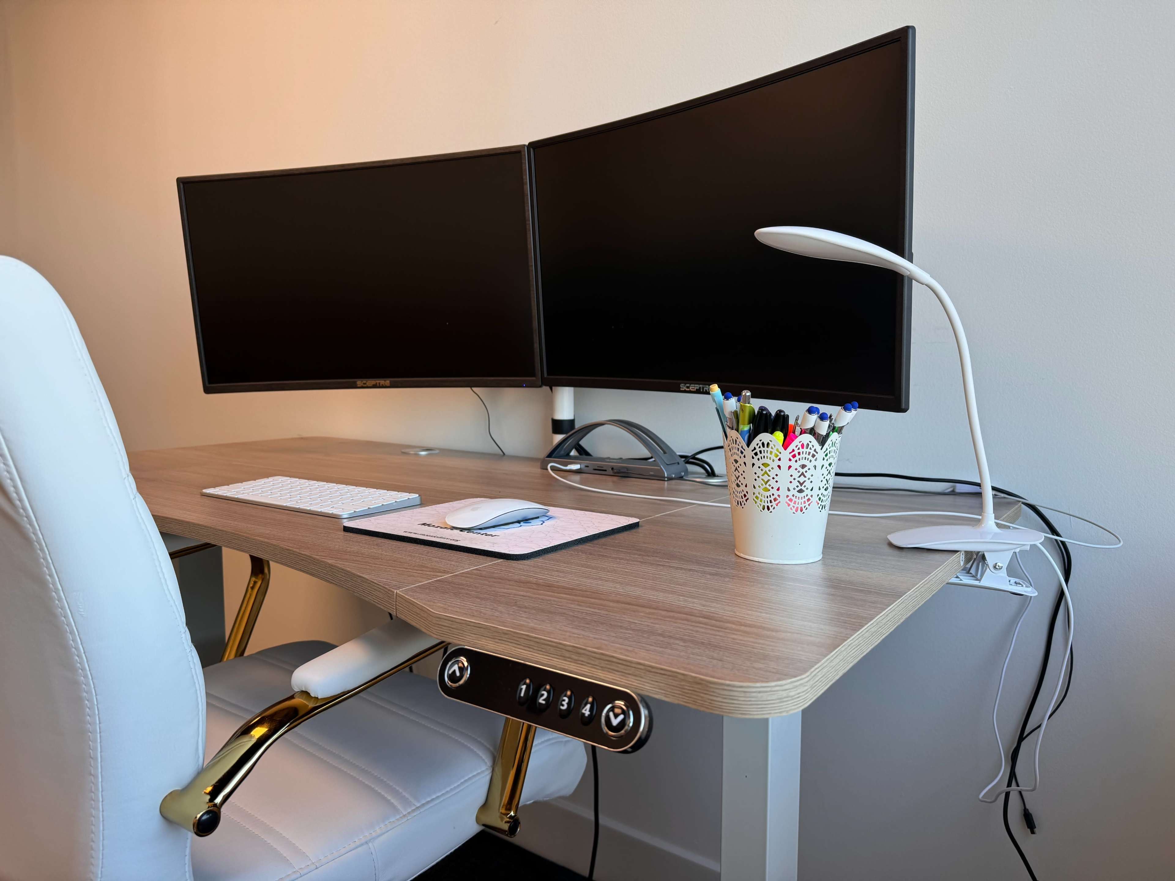A modern desk setup features two curved monitors, a keyboard and mouse, a decorative pen holder, and a task lamp.