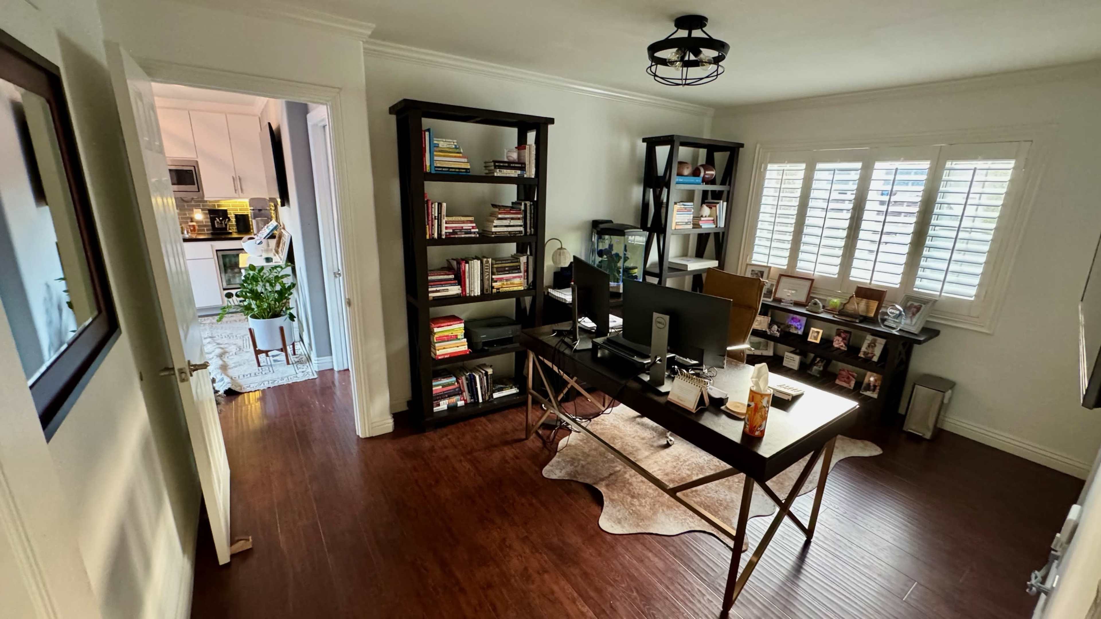 The image shows a home office with a desk and two shelves filled with books, framed photos, and a plant visible through an open doorway.