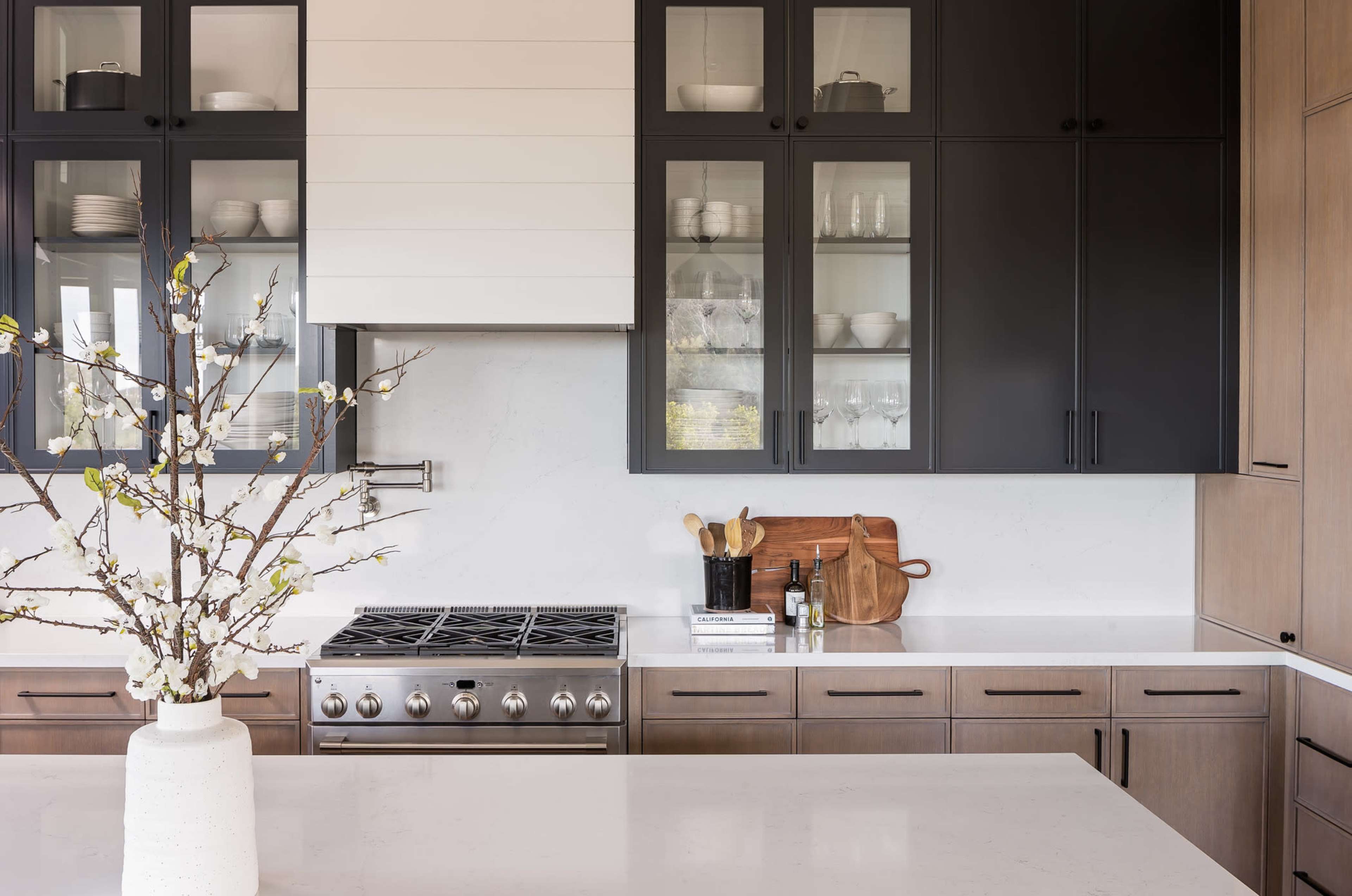 A modern kitchen featuring dark cabinetry, a marble countertop, and glass-fronted upper cabinets displaying dishware, with a vase of branches on the counter.