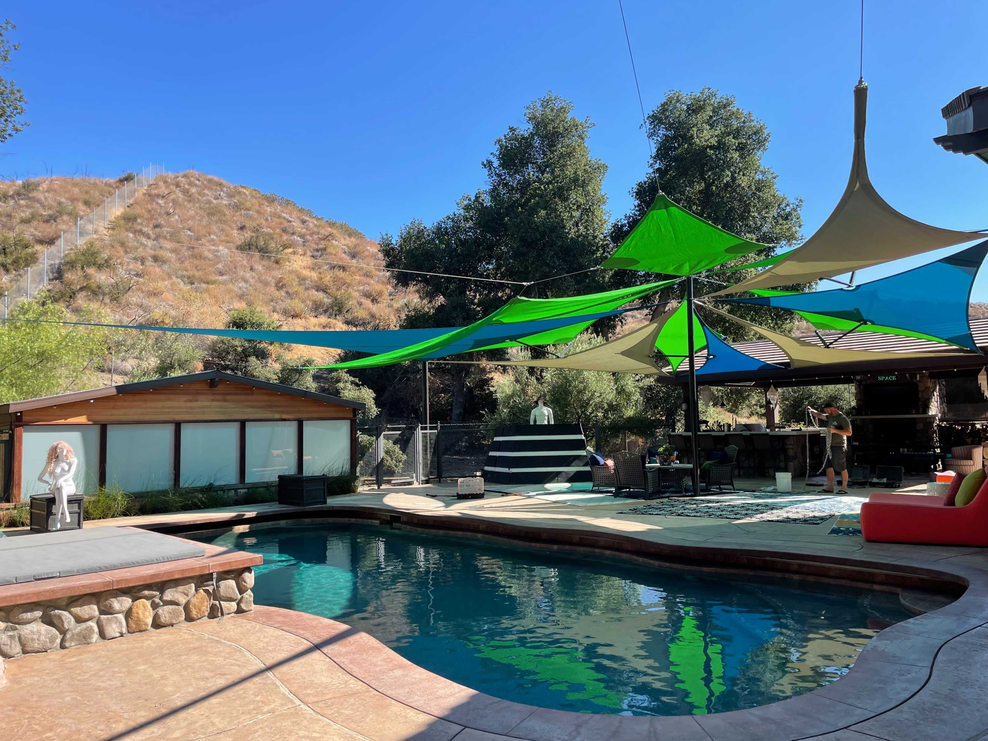 A swimming pool area features colorful fabric shades overhead, surrounded by green trees and a hillside in the background.