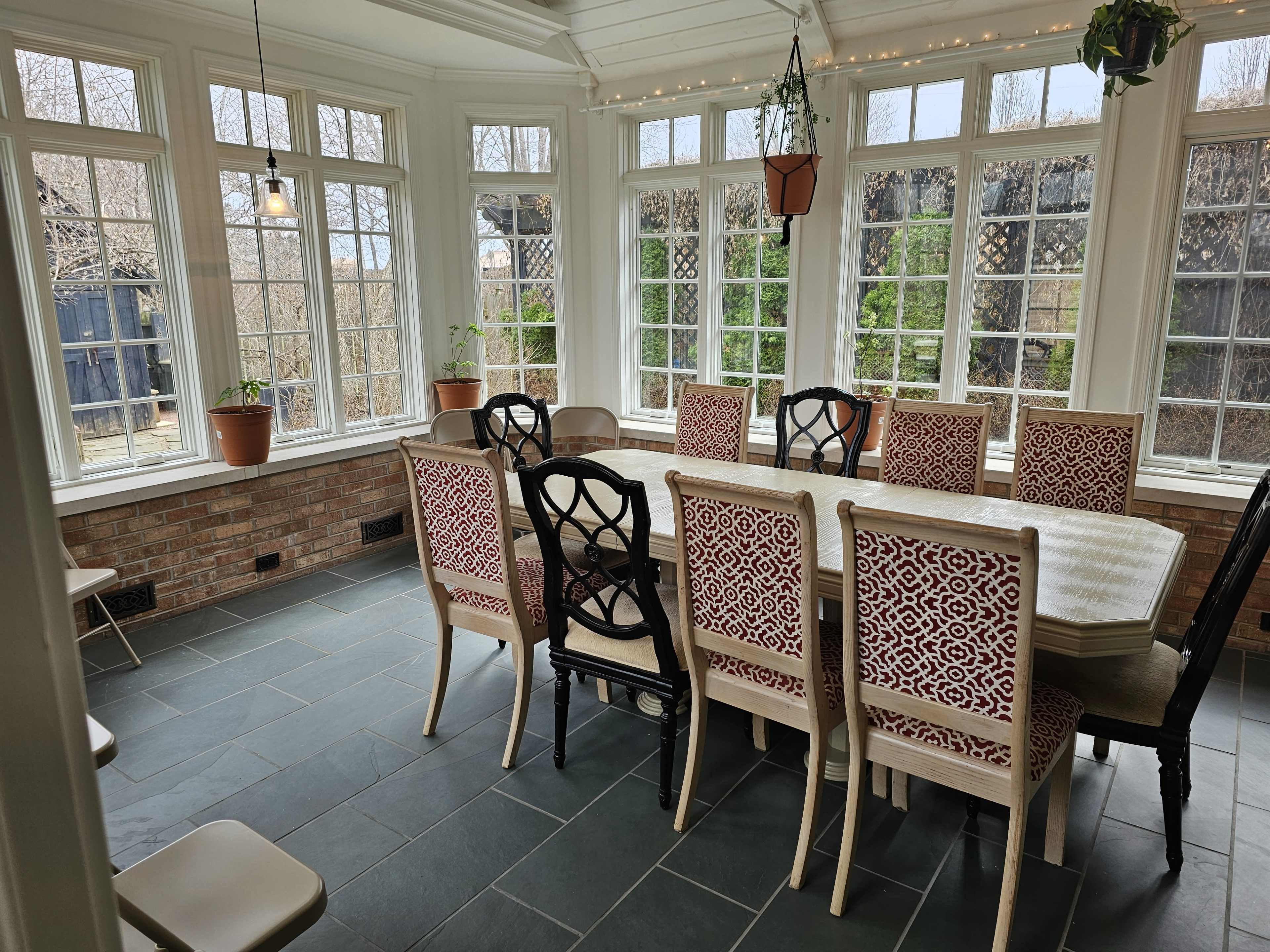 A sunlit dining area features a large, rectangular table surrounded by patterned chairs, with windows providing views of greenery outside.