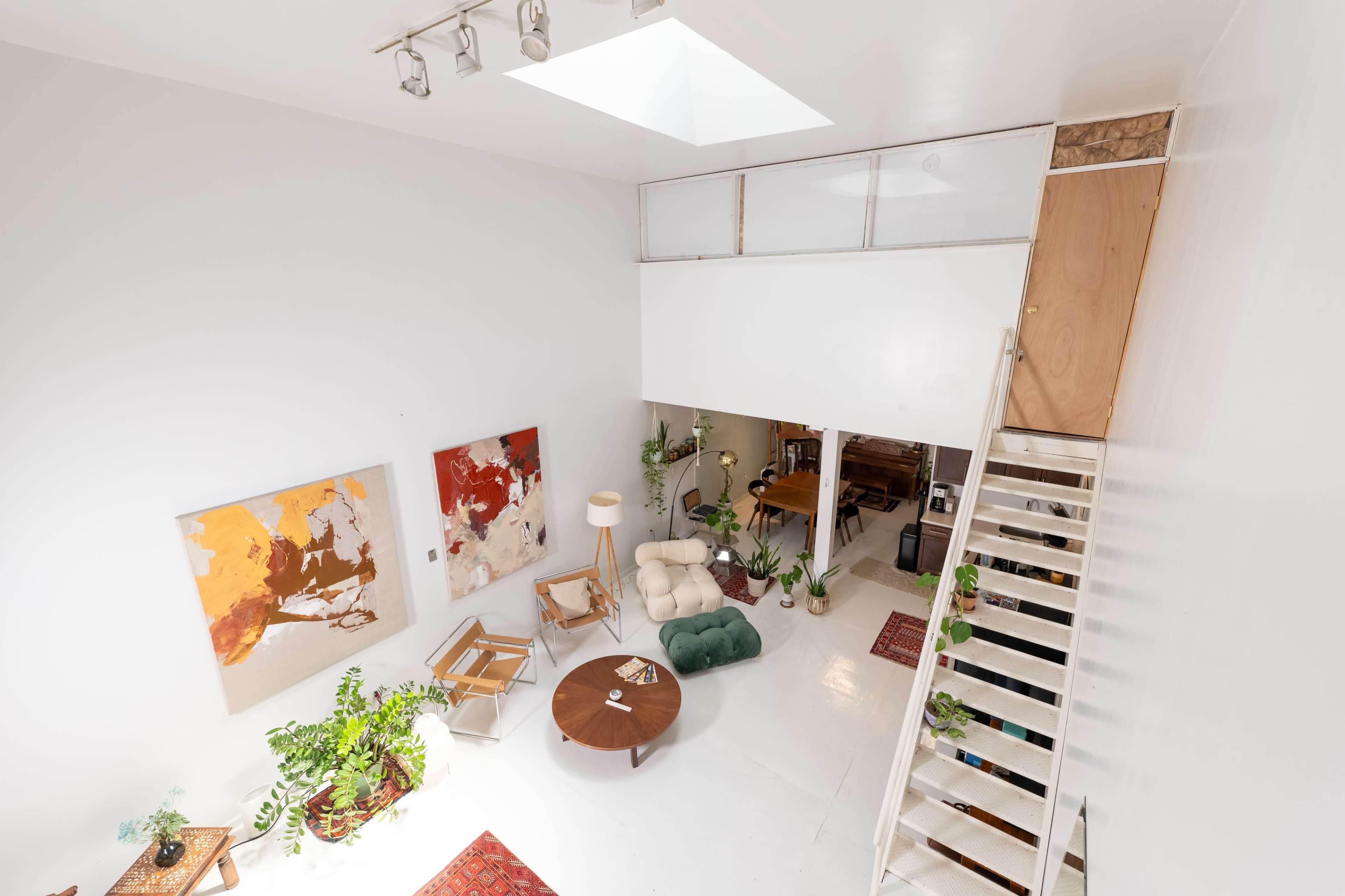 A bright, open living space features a staircase, a round coffee table, and several plants, with artwork displayed on the white walls.