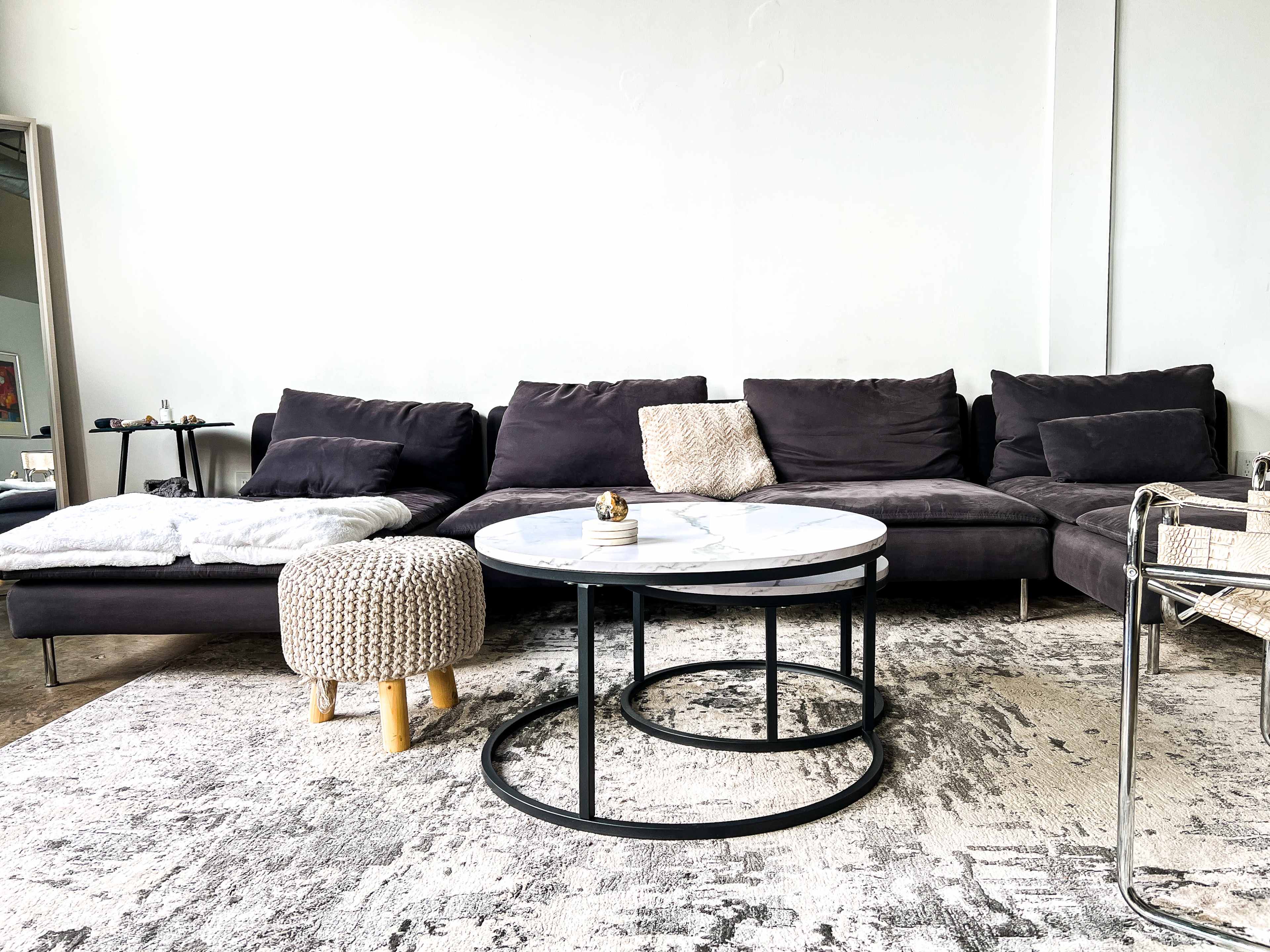A modern living room features a dark gray sectional sofa, a round marble coffee table with metal legs, and a light-colored knitted pouf on a large area rug.