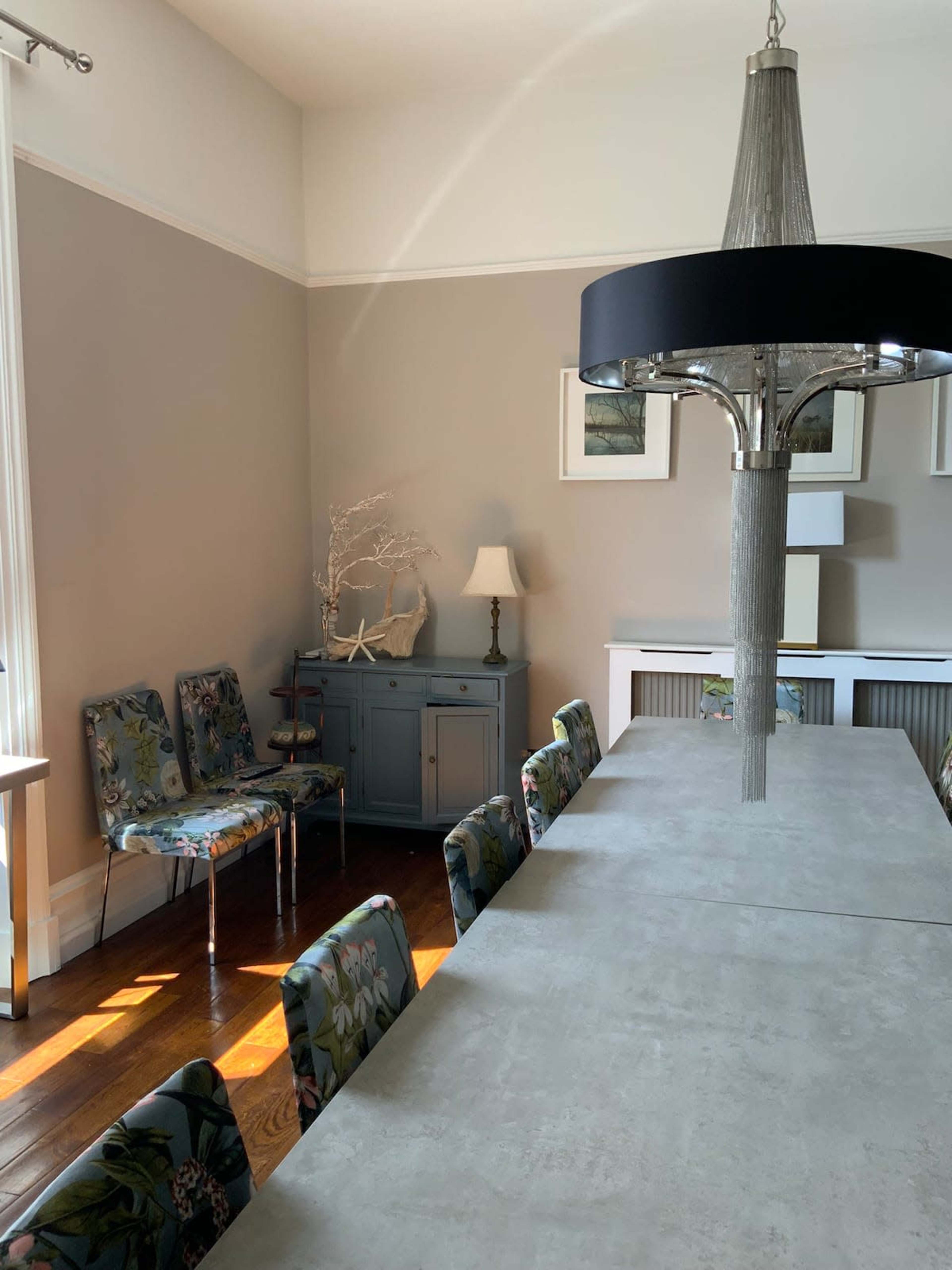 A modern dining space features a long table with floral upholstered chairs, a pendant light, and a side table alongside a lamp.