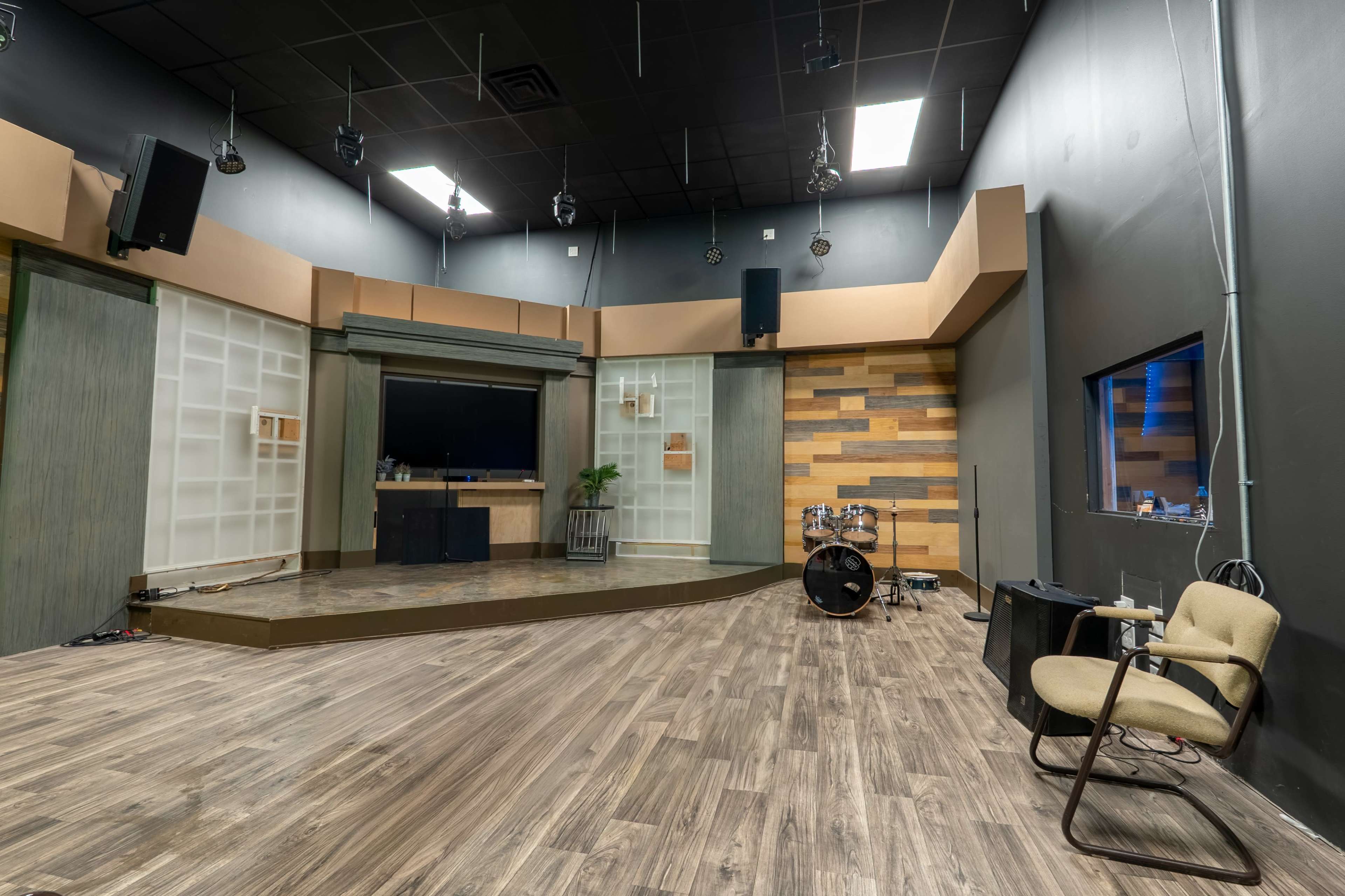 The image shows a spacious studio with wooden flooring, featuring a stage area, a television screen, and various sound equipment.