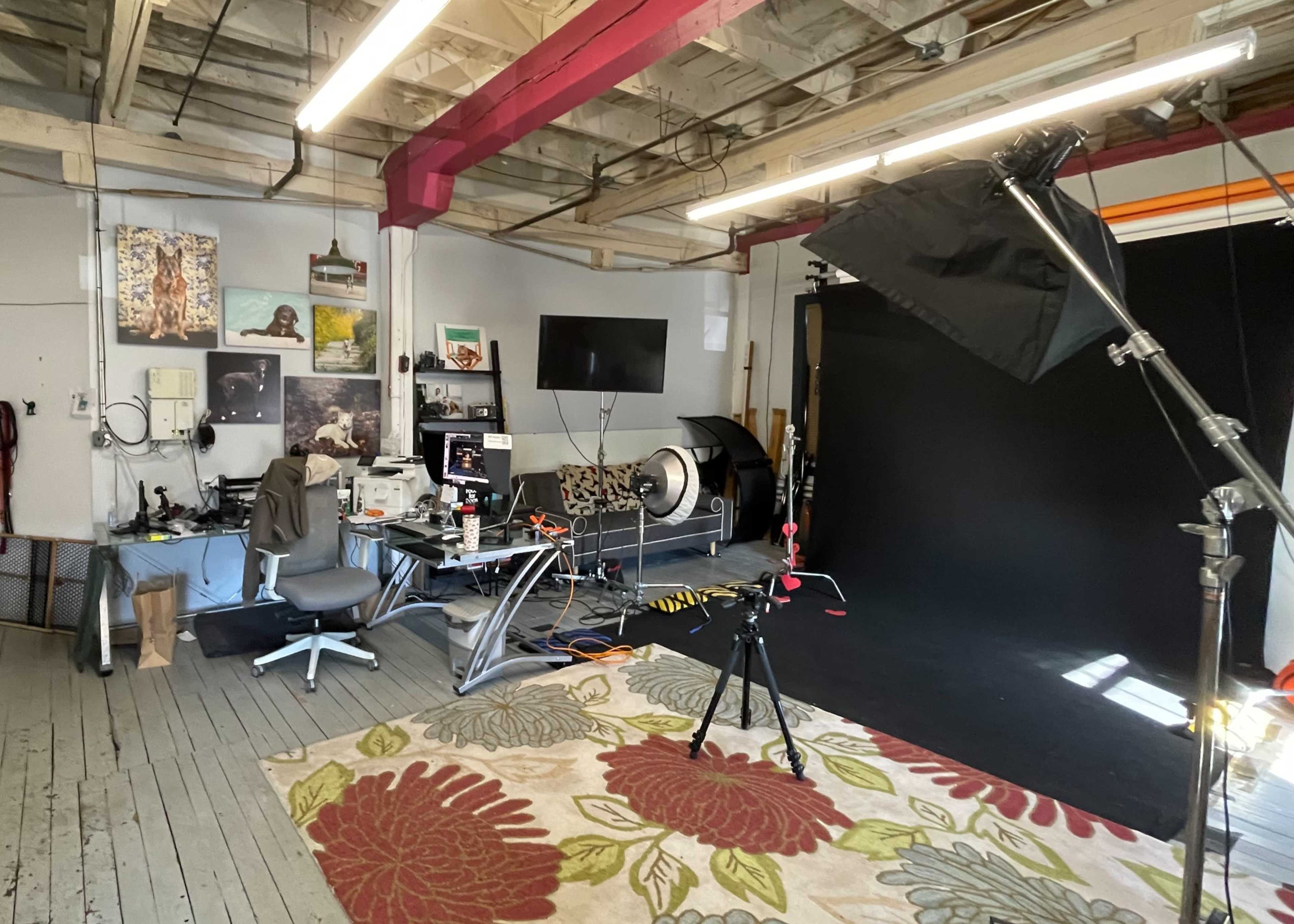 A photography studio with a desk, computer equipment, and a large black backdrop set up for shooting.