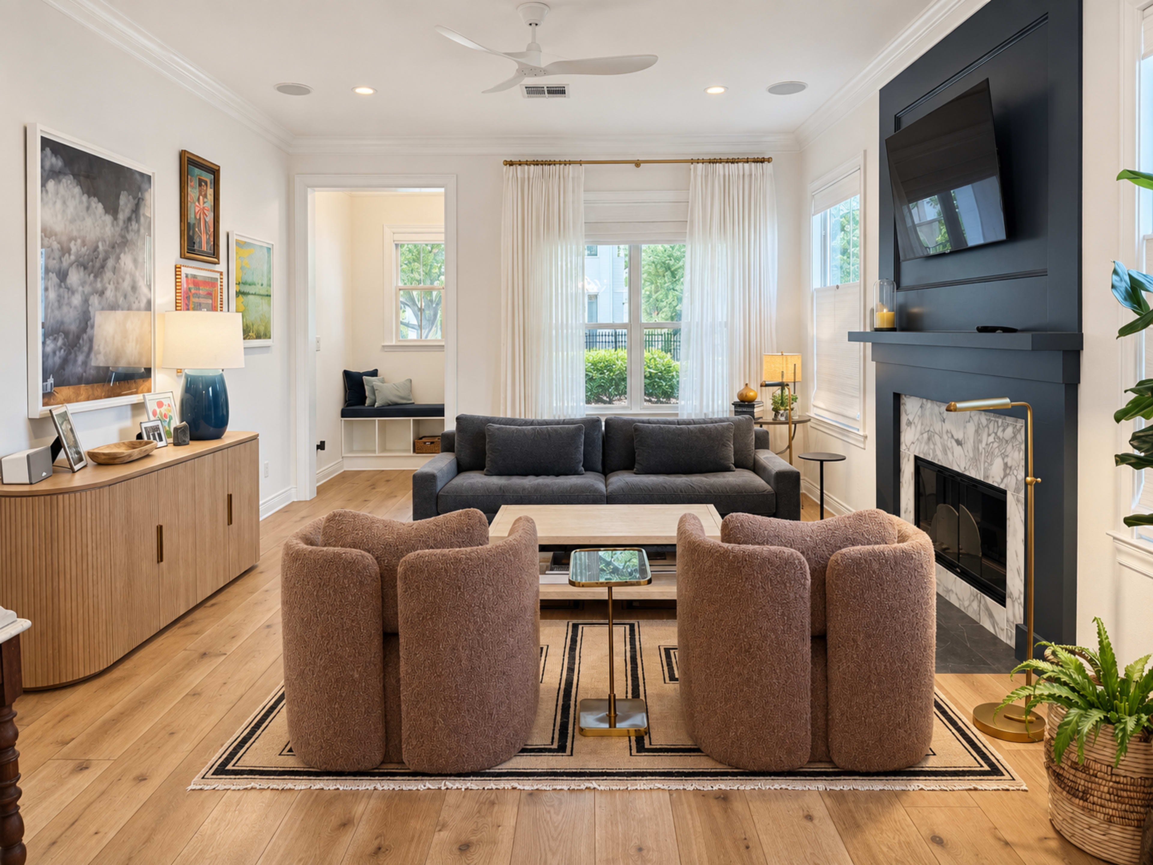 The image shows a modern living room featuring a dark couch, two rounded armchairs, a wooden sideboard, and a fireplace with a TV mounted above it.