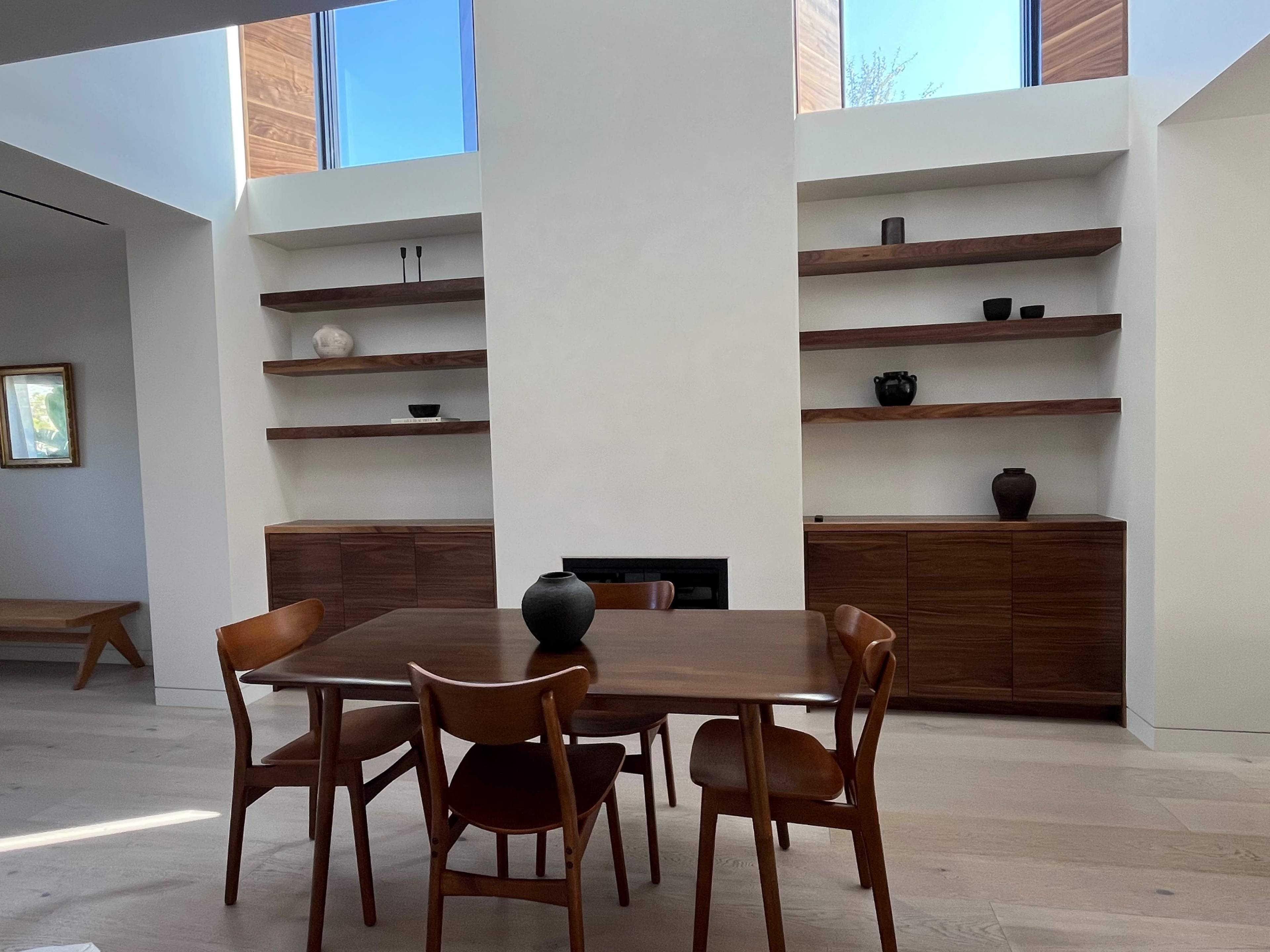 A modern dining area features a wooden table surrounded by four chairs, with open shelves and cabinets against a light-colored wall.