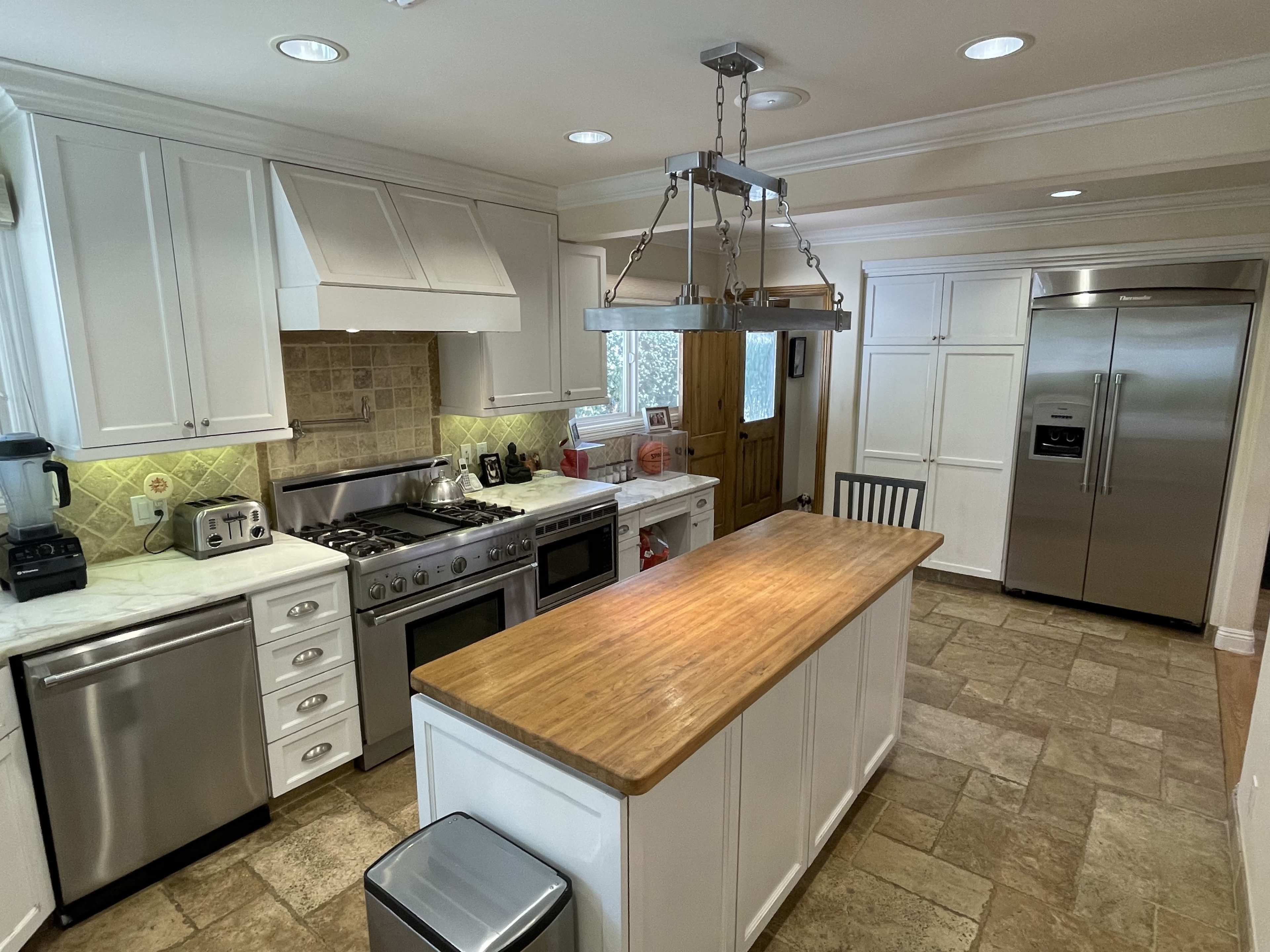 A kitchen with white cabinets, a wood-topped island in the center, stainless steel appliances, and a large refrigerator.