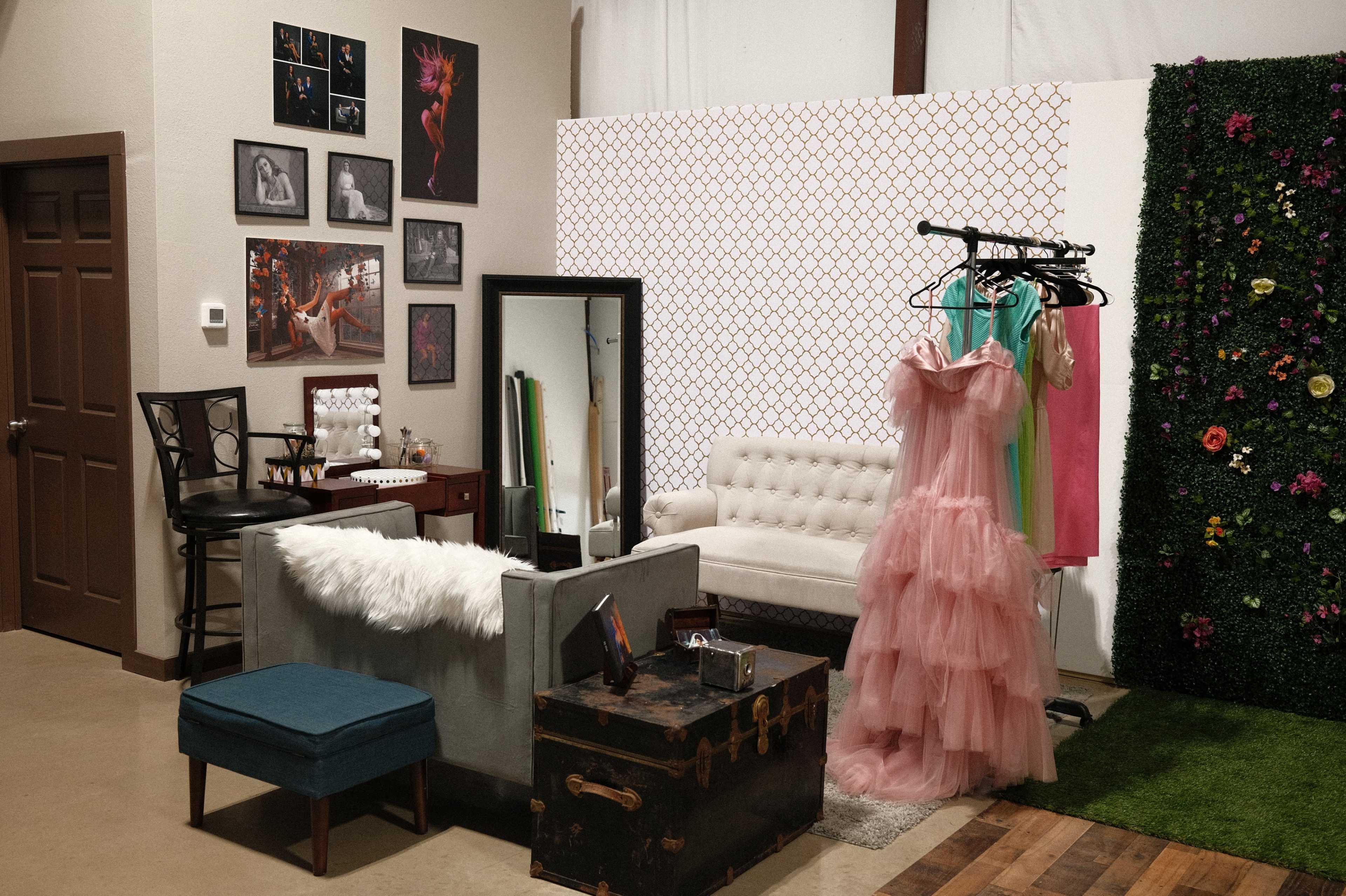 A styled interior space with a vintage sofa, a mirror, a clothing rack with colorful dresses, and a wall adorned with framed art and greenery.