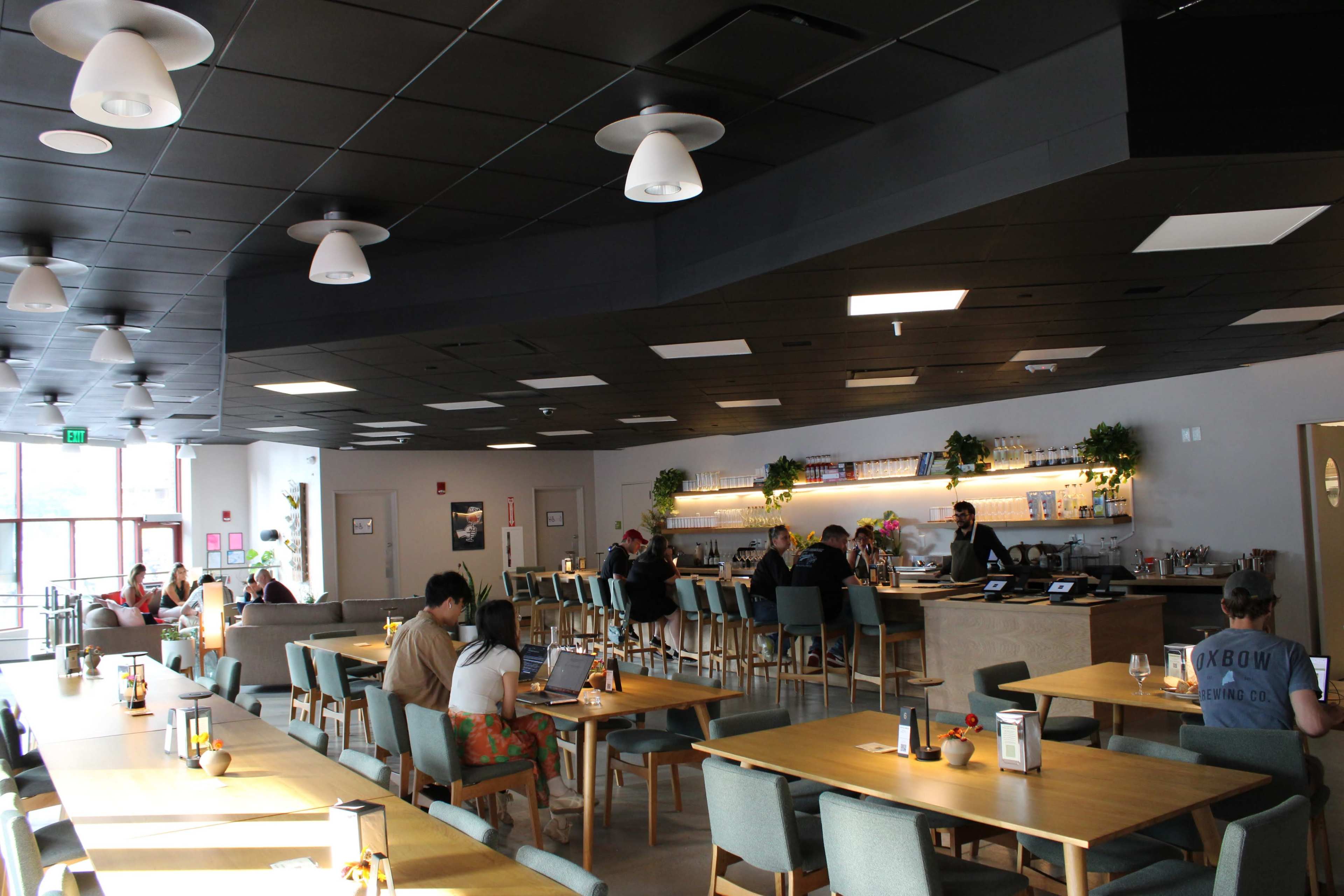 A modern café features several tables occupied by customers working on laptops and enjoying their meals, with a long counter displaying food and beverages in the background.