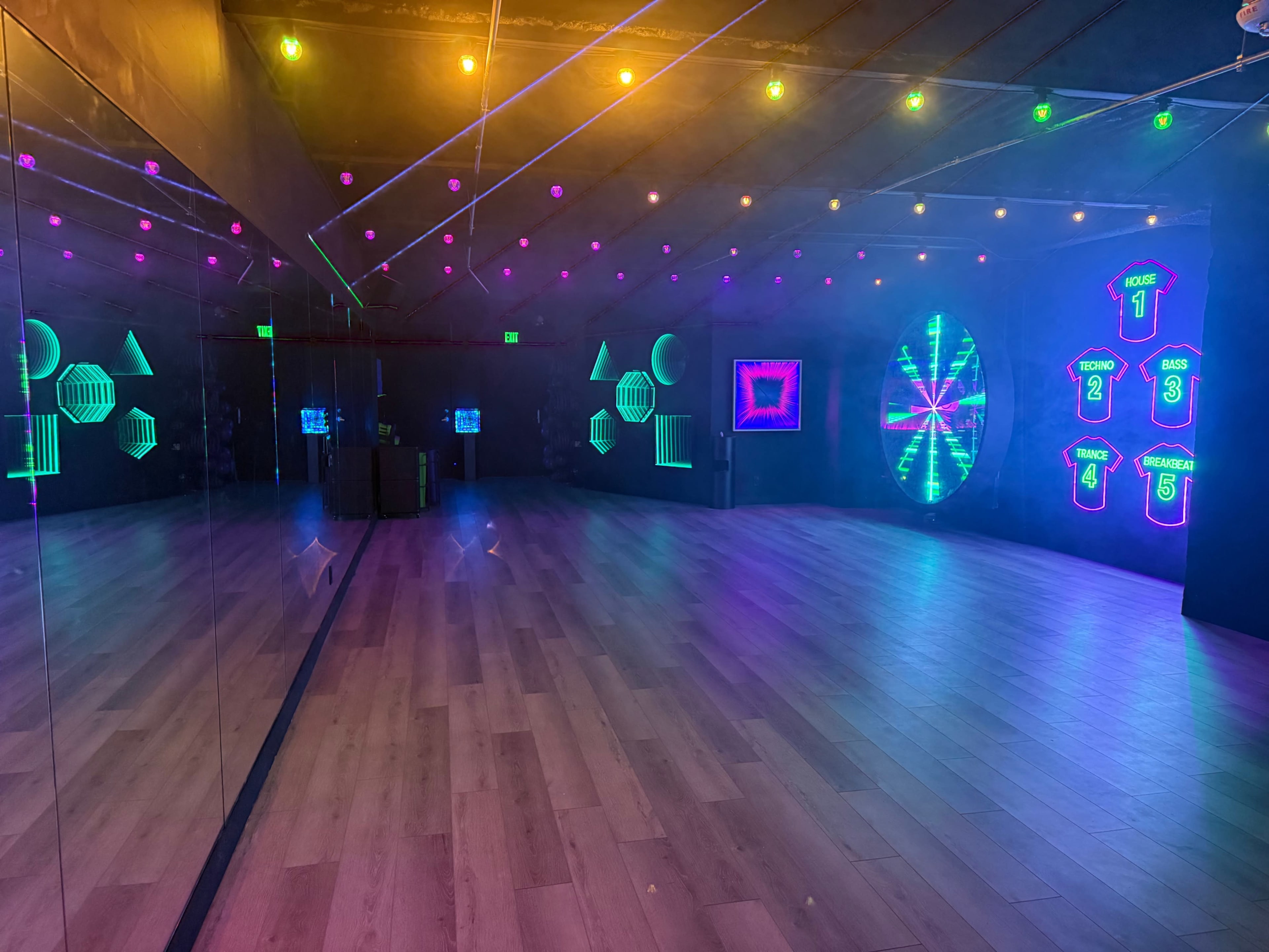 The image shows a dimly lit room with colorful neon lights and geometric shapes displayed on the walls, along with a mirrored surface and a light-up wheel in the center.