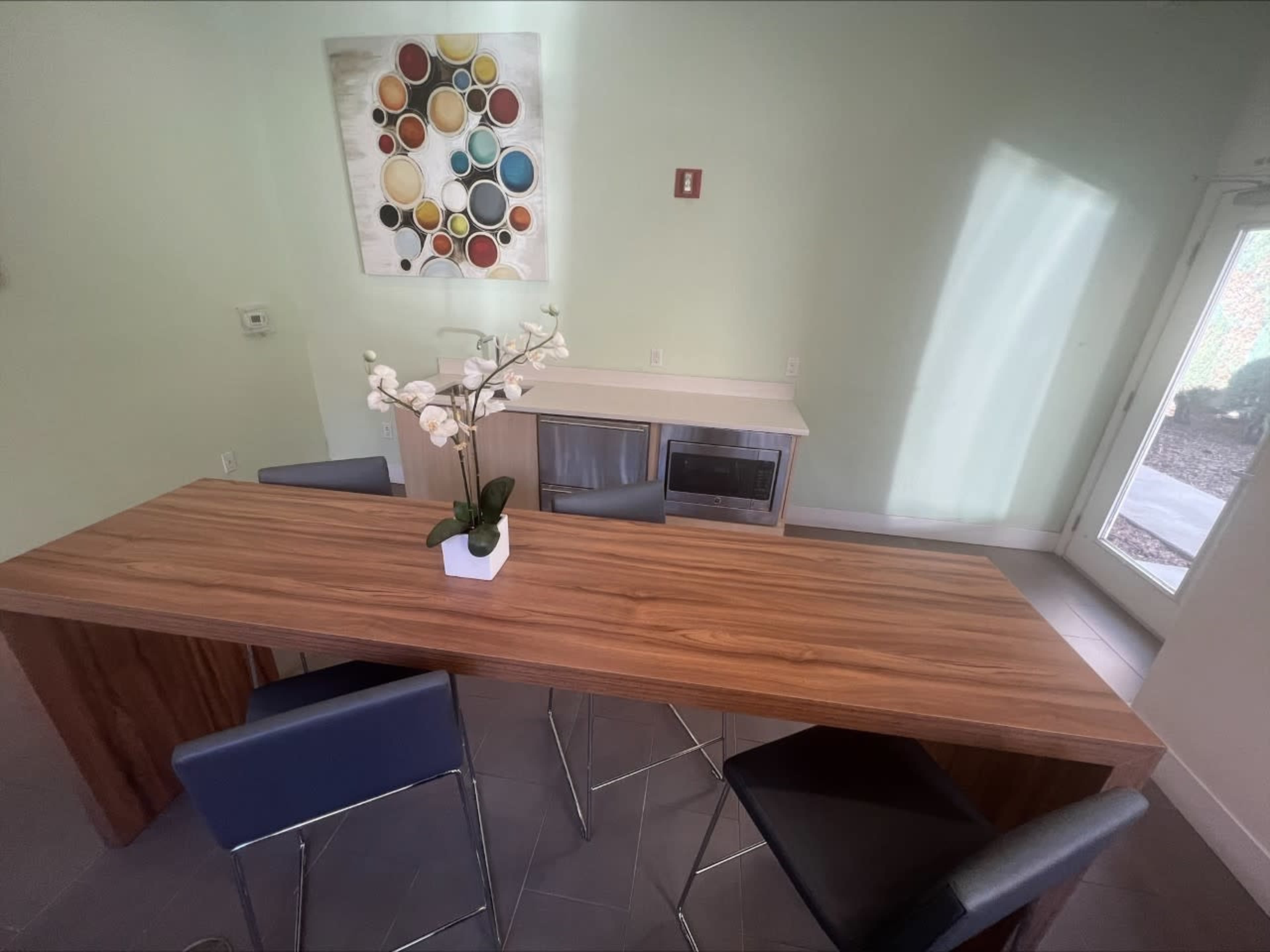 A wooden dining table with black chairs is positioned in a light-colored room, featuring a cabinet and a decorative wall piece above.