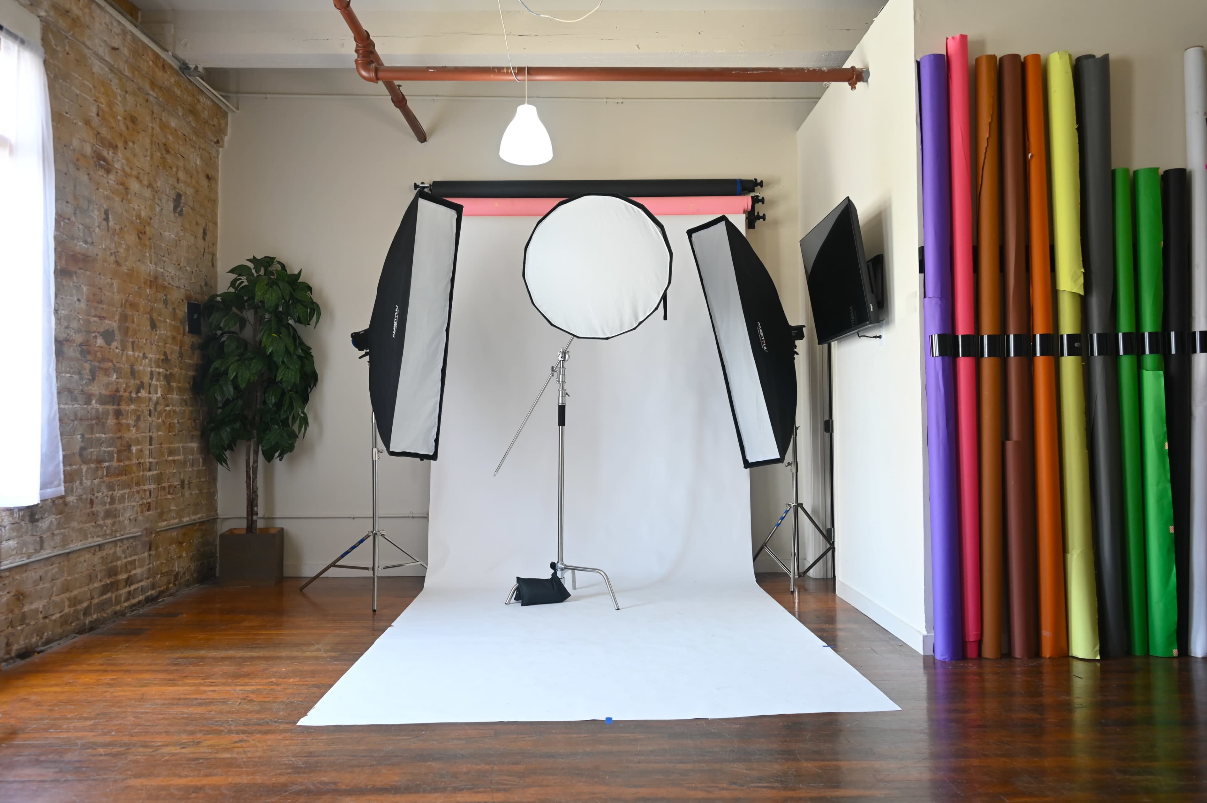 A photography studio with softbox lighting setups, a white backdrop, and a collection of colorful backdrop rolls on one side.