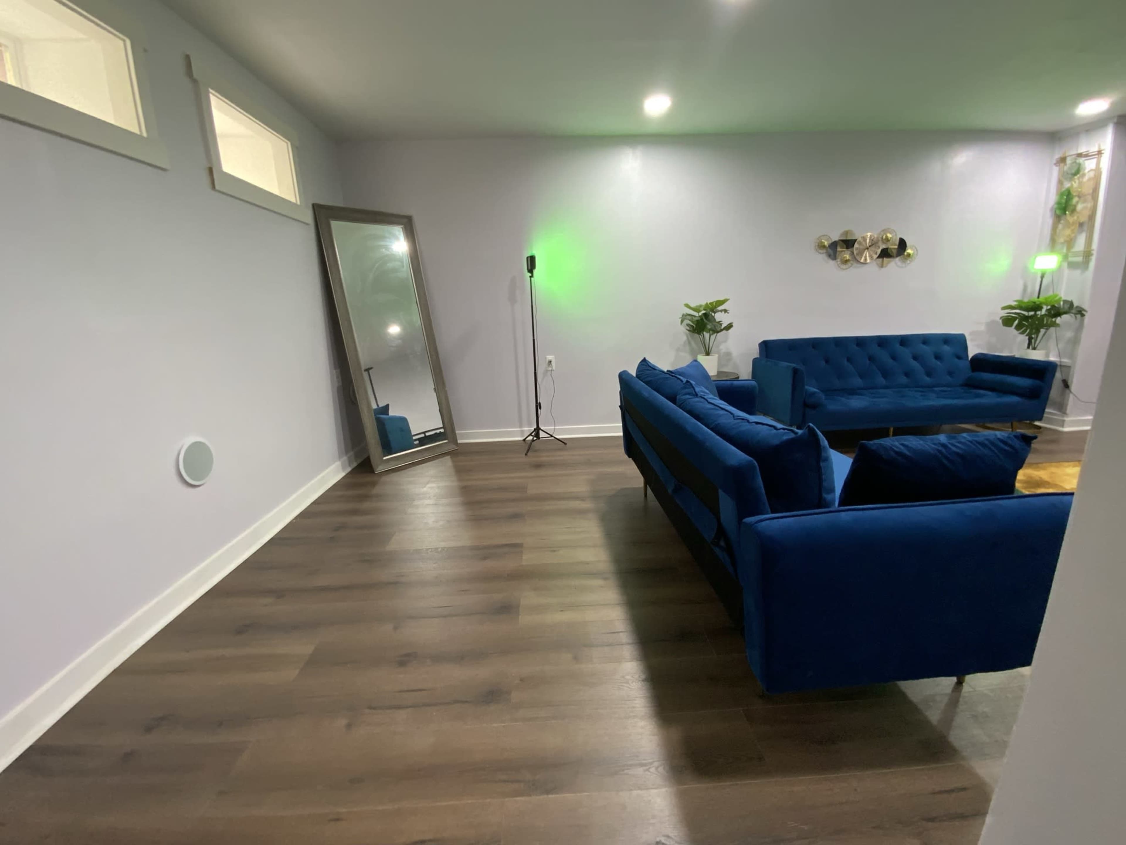 A modern living room featuring a blue sofa, a large mirror, green ambient lighting, and hardwood floors.