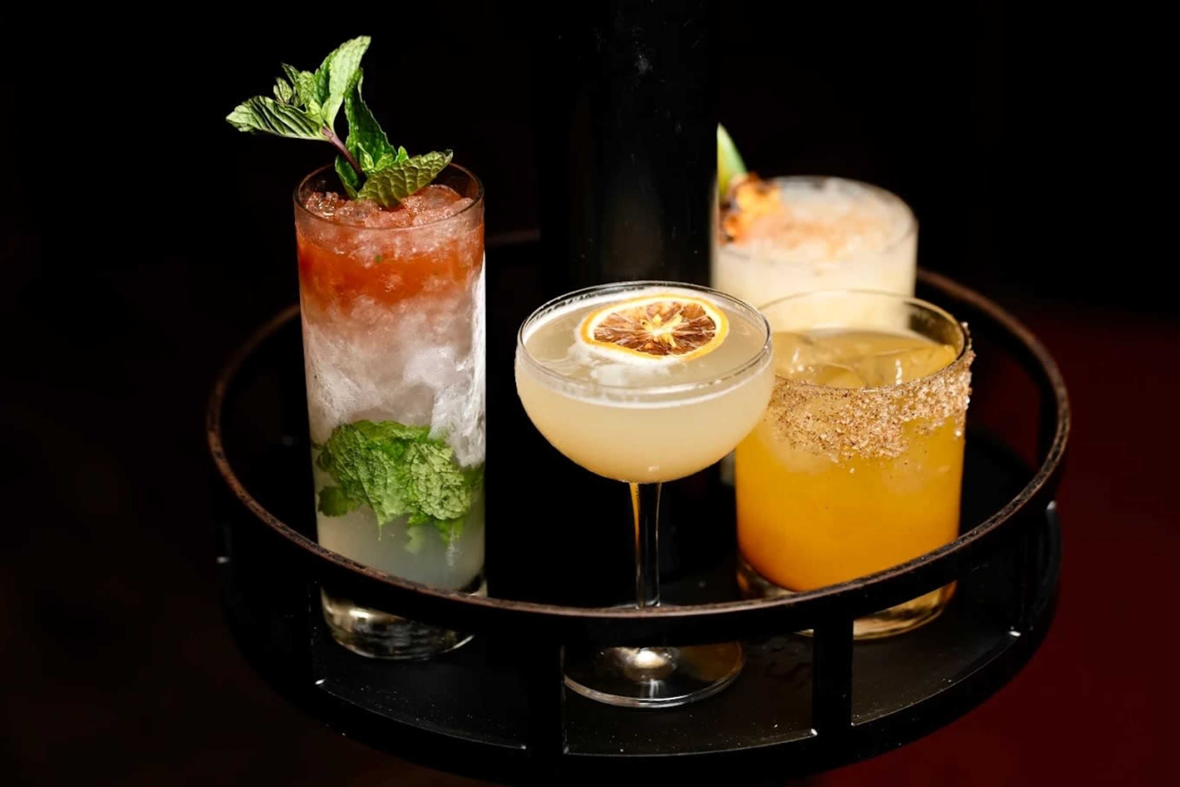 The image features a tray holding four distinct cocktails, each presented in different glassware with garnishes such as mint, dried fruit, and sugar rim.