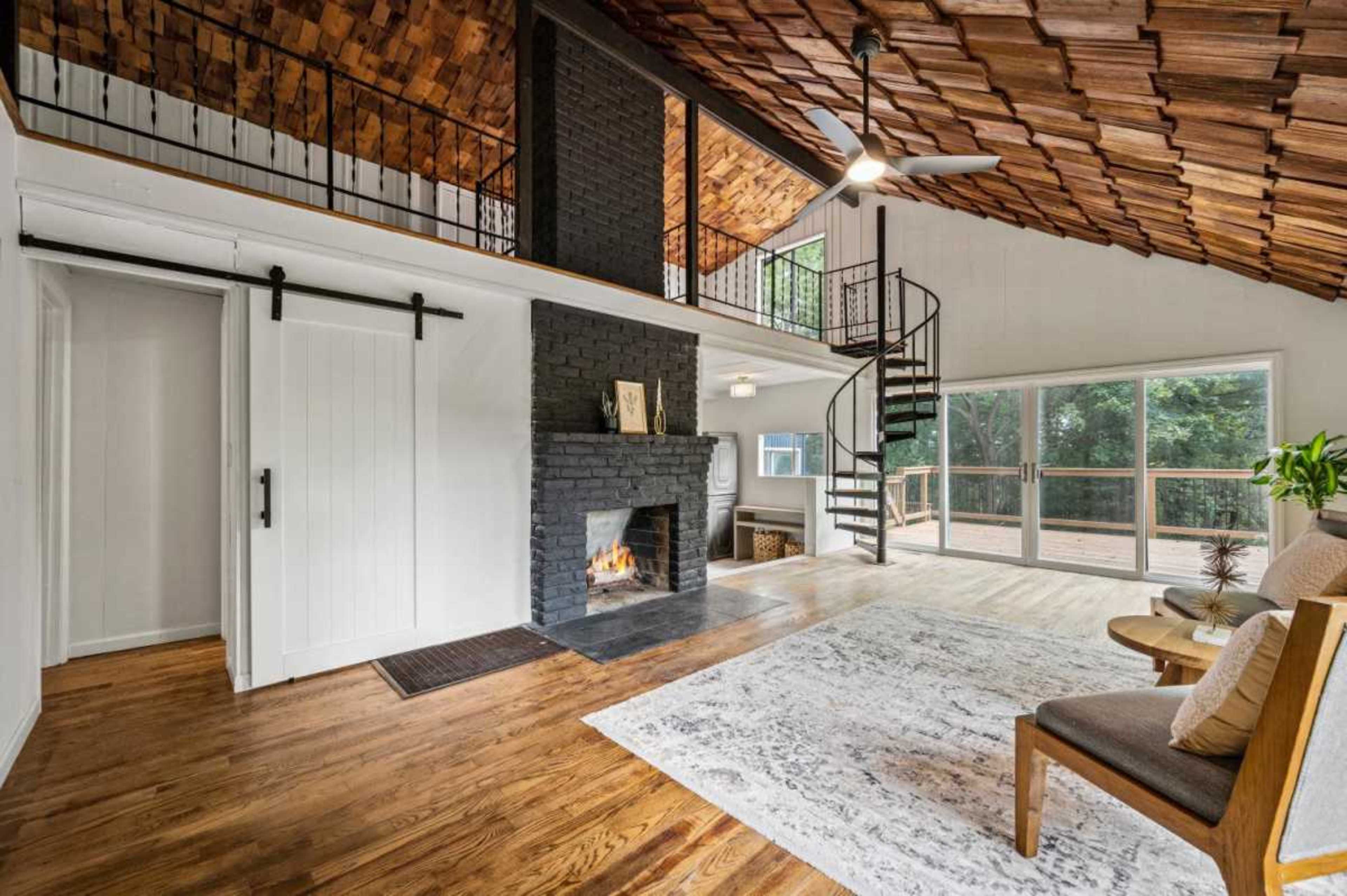 The image shows a cozy living area with a wood-paneled ceiling, a black stone fireplace, a spiral staircase, and large glass doors leading to an outdoor space.