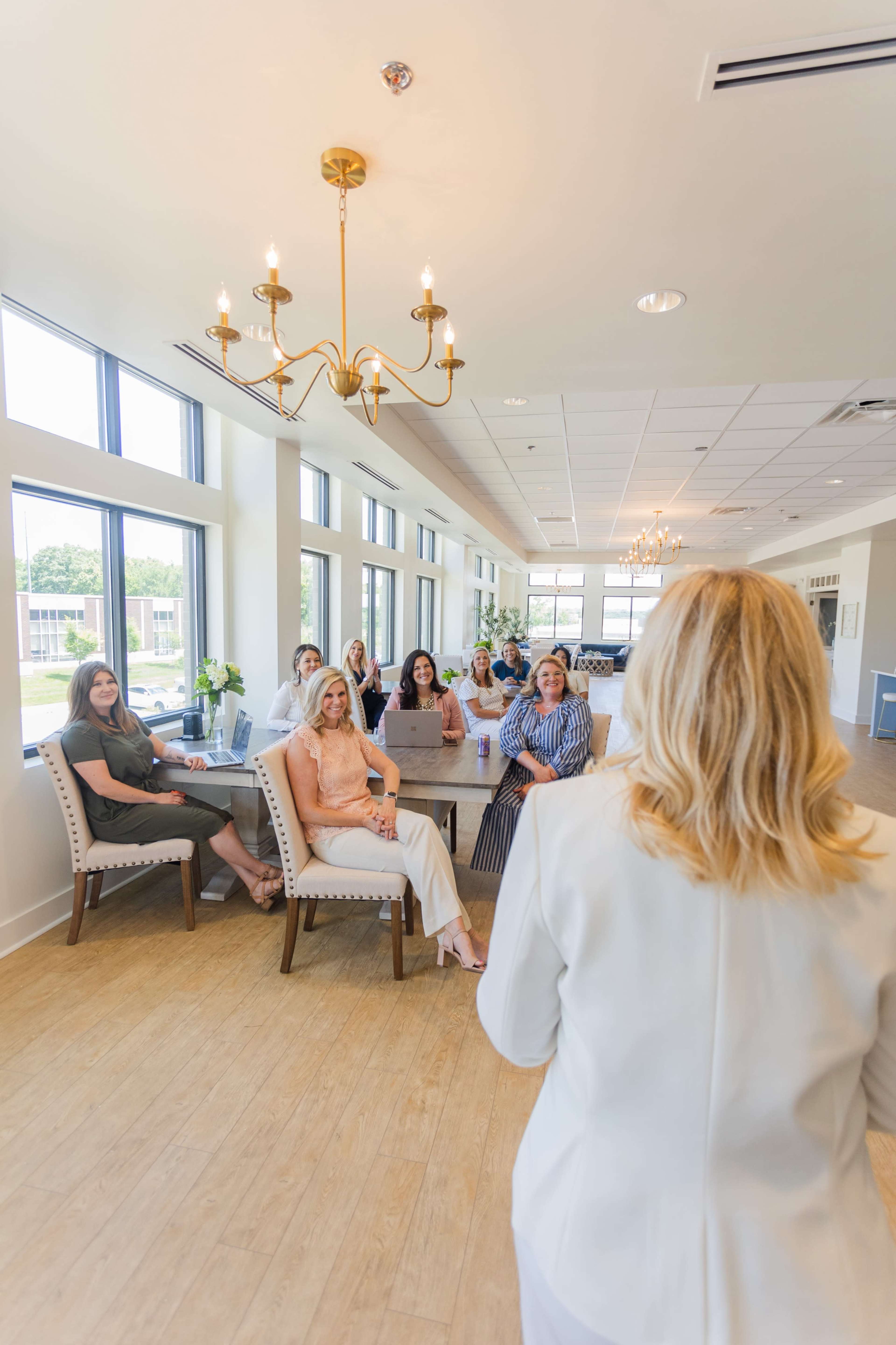 A group of seven women sits in an elegantly designed room with large windows, while one woman stands in front, addressing the others.