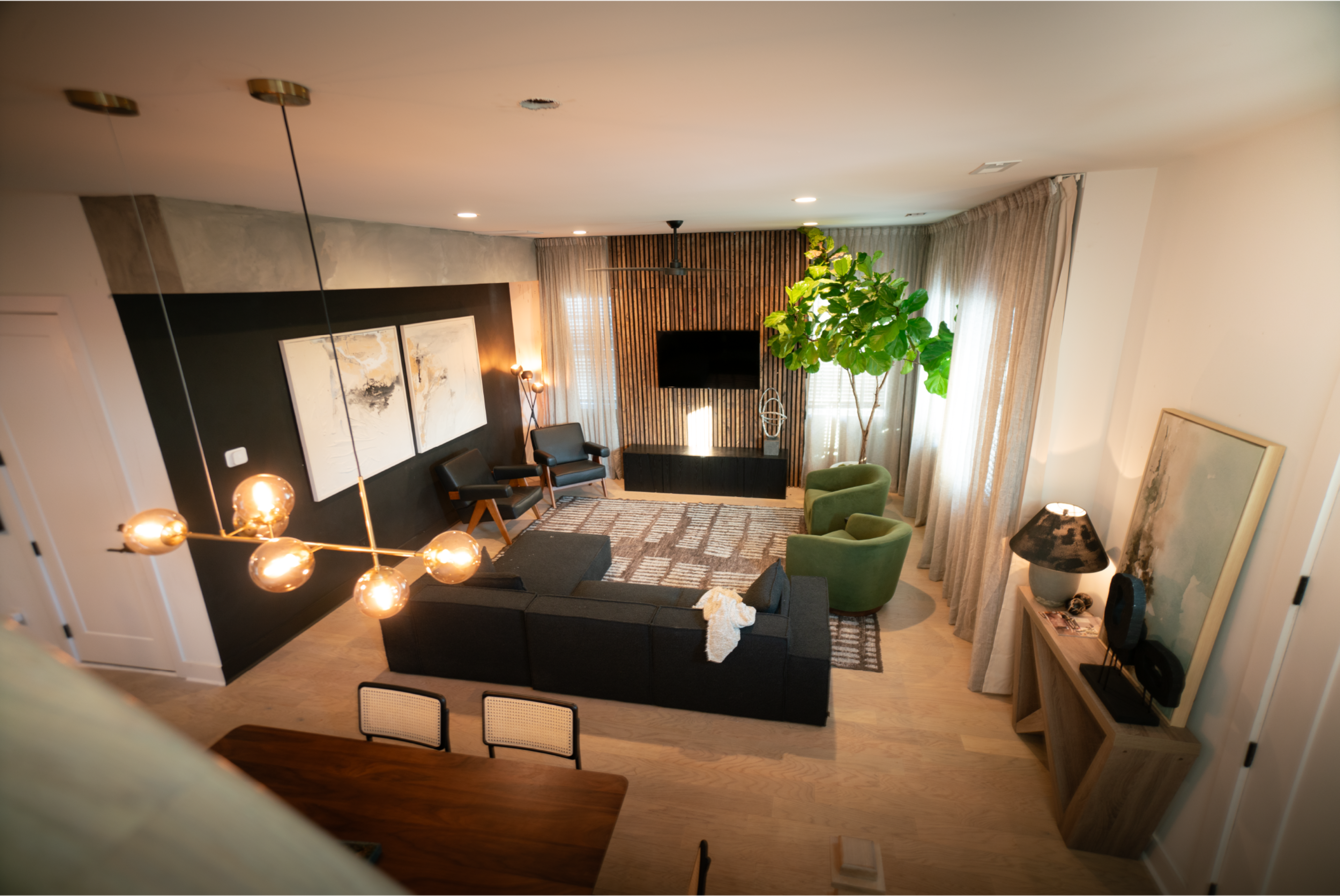A modern living room featuring a sectional sofa, two green armchairs, a large television, and large windows with sheer curtains.