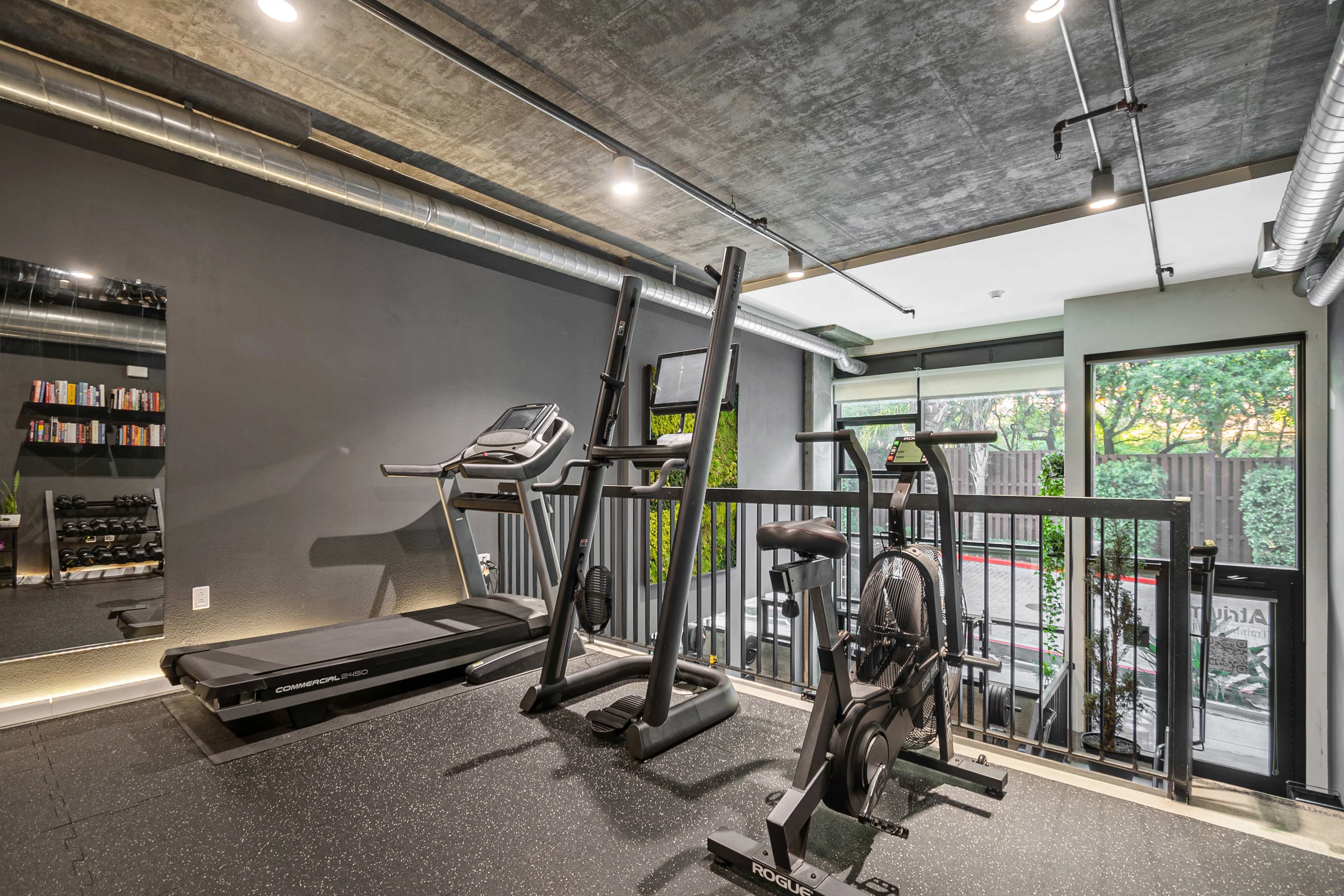 A modern fitness area featuring a treadmill, an elliptical machine, and a stationary bike, with large windows and a polished concrete ceiling.