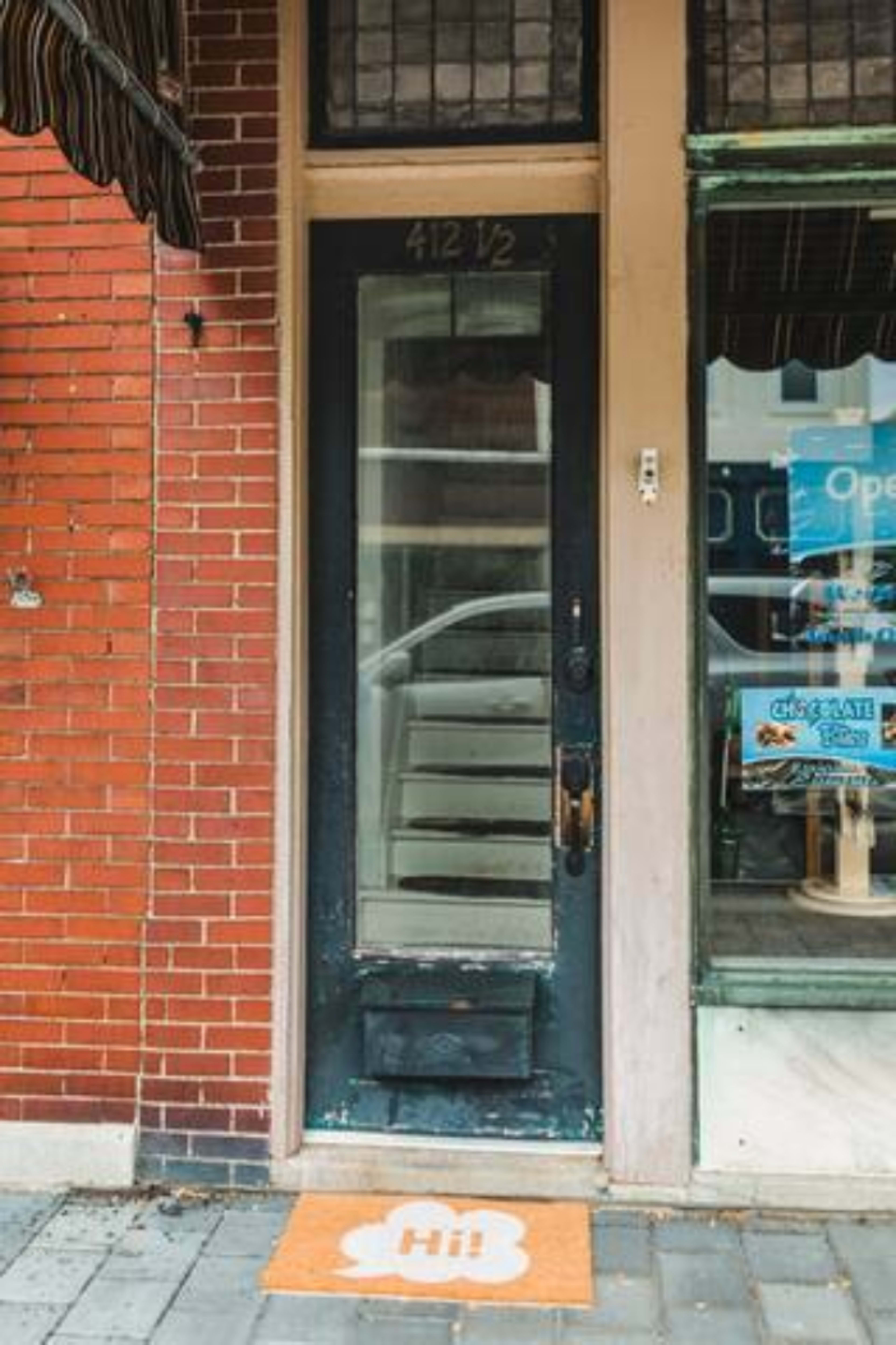 The image shows a black door with a glass panel and a welcome mat in front, located in a brick building with a store window nearby.