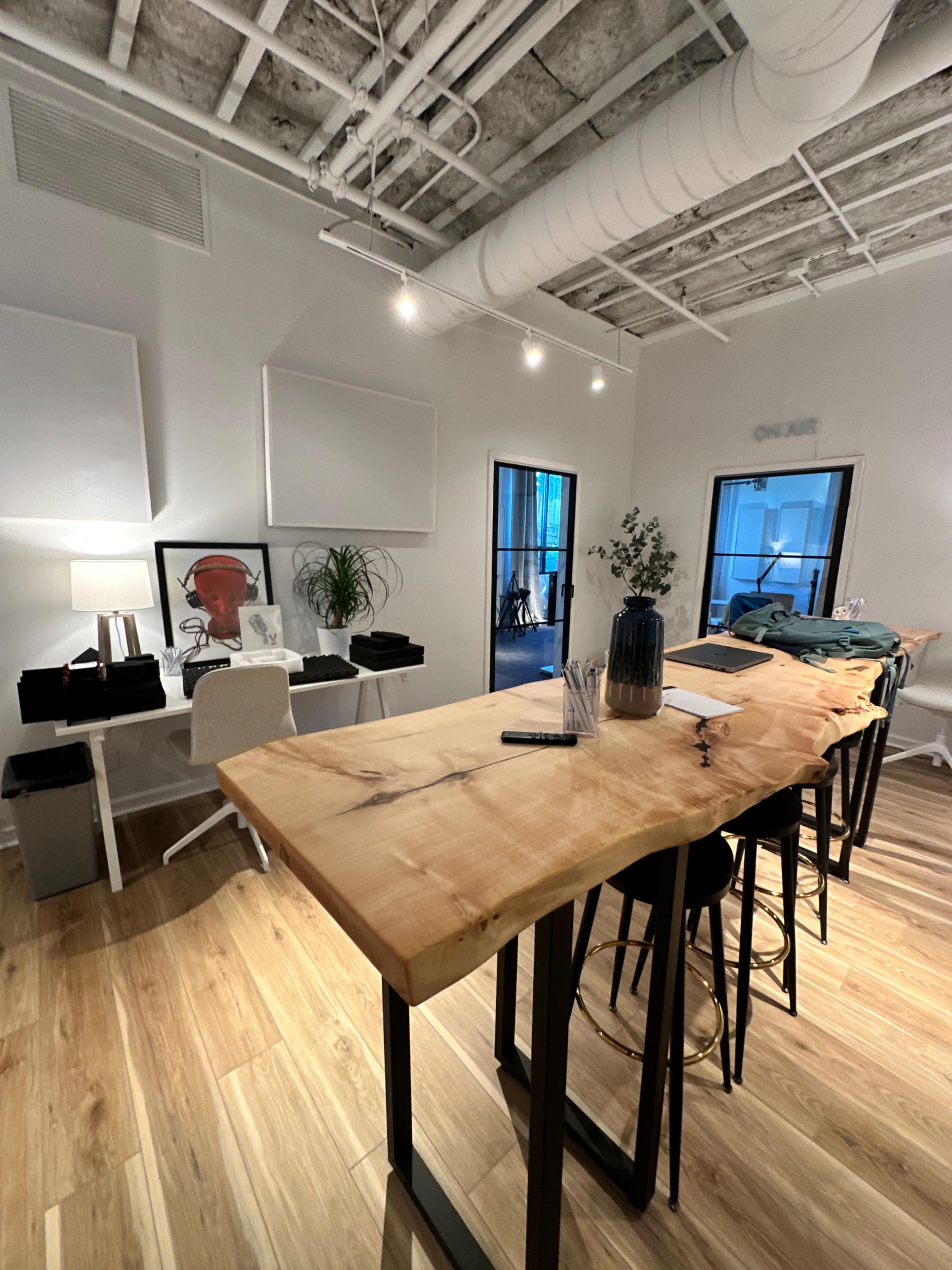 The image shows a modern office space featuring a large wooden table with stools, surrounded by minimalist decor, plants, and two glass doors leading to another room.