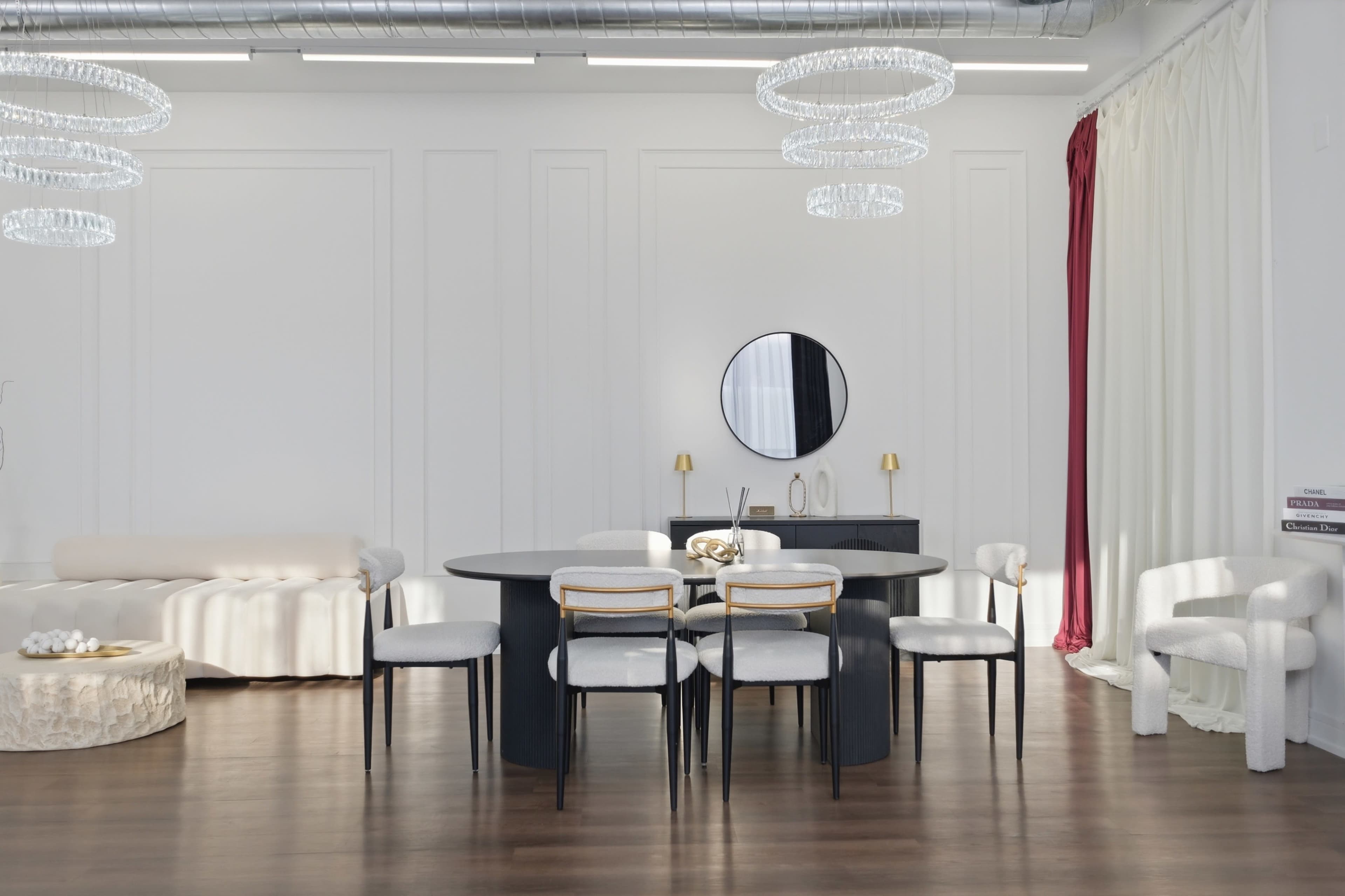 The image shows a modern dining area featuring a round black table surrounded by six chairs, with a minimalist white and gray color scheme and two contemporary light fixtures hanging from the ceiling.