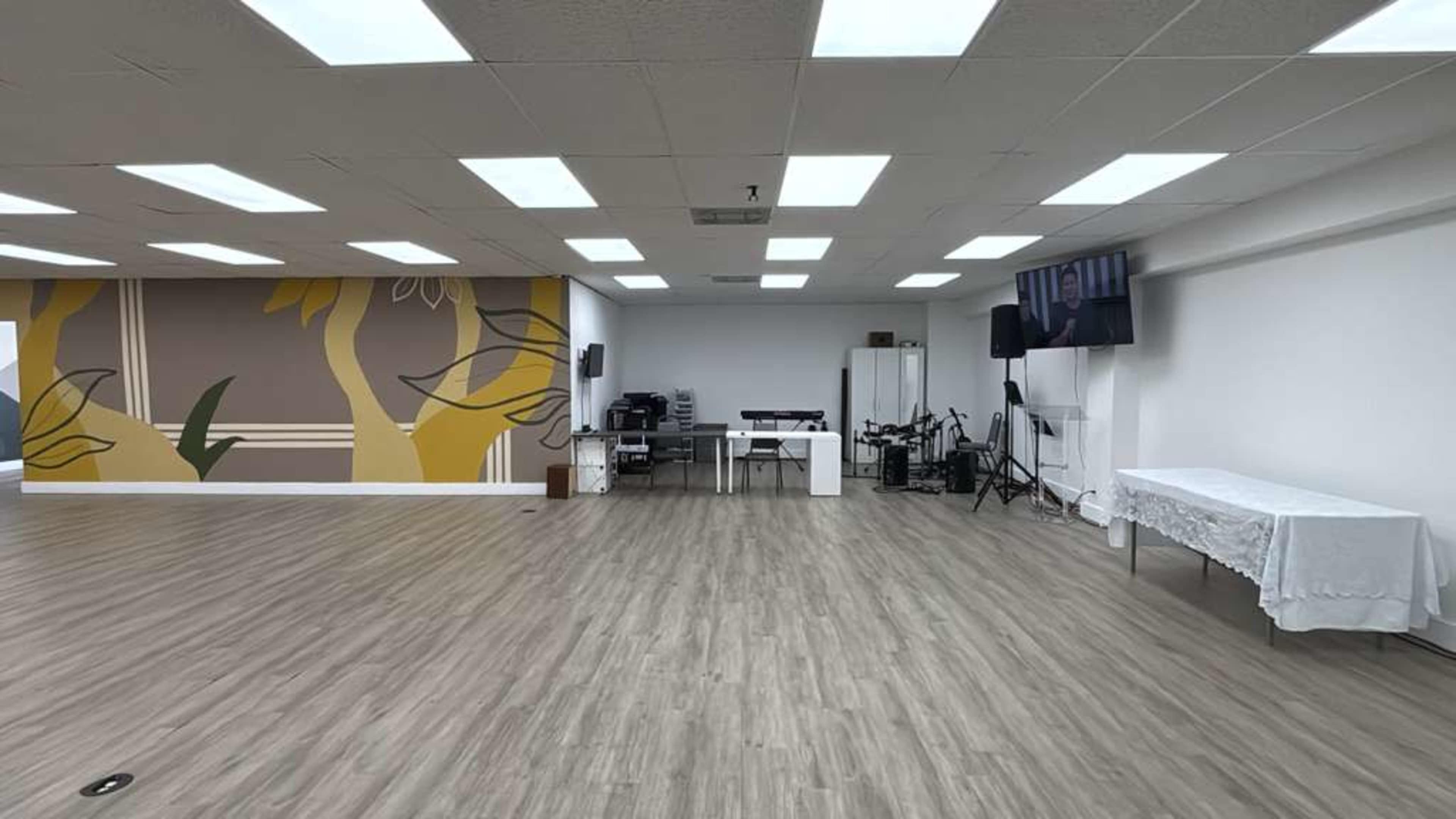 The image shows a spacious and well-lit room with a wooden floor, featuring a mural on one wall, a table covered with a white cloth, and electronic equipment set up in the background.
