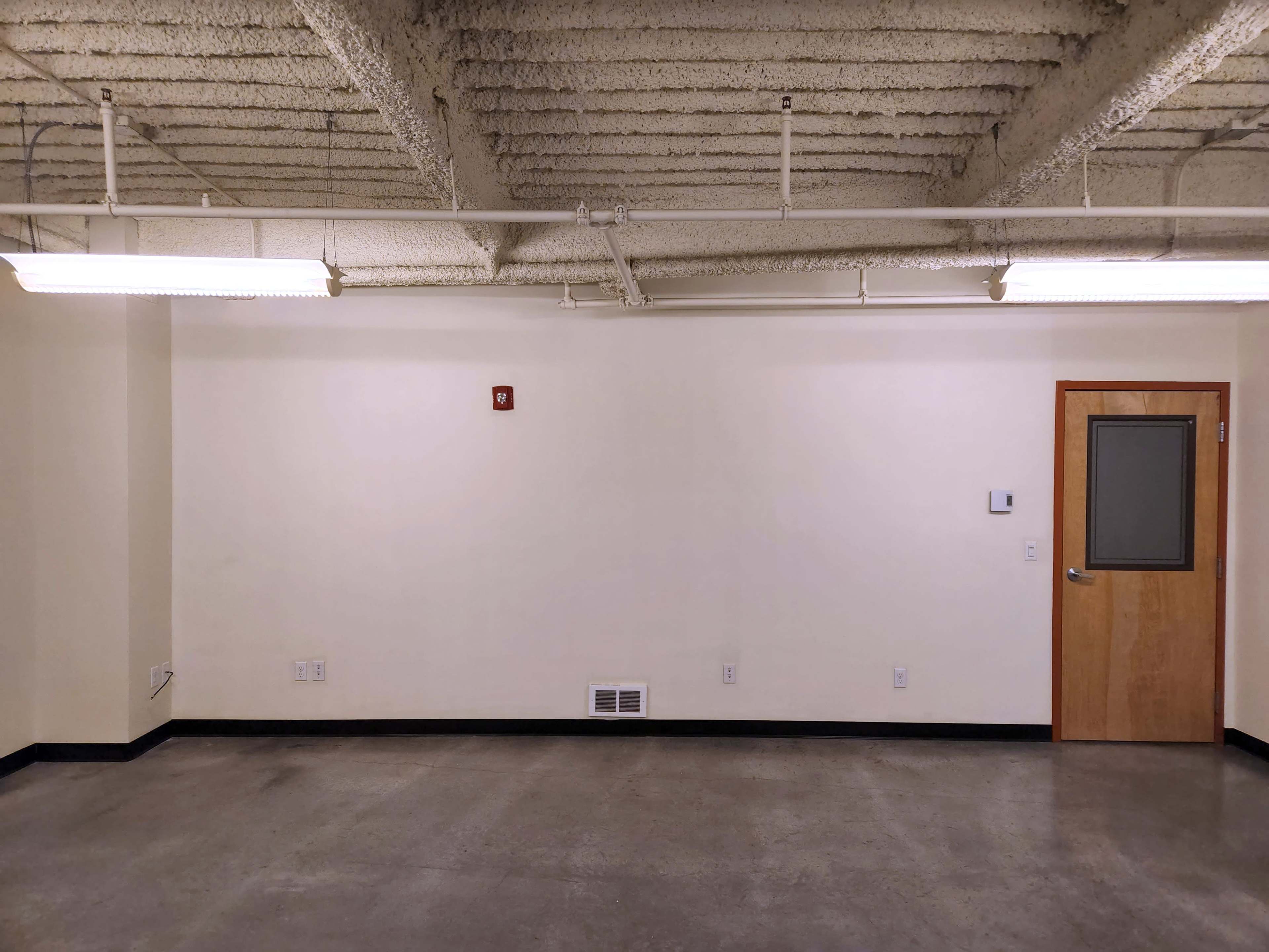 A blank wall with a door and minimal fixtures in an empty room featuring concrete flooring and exposed ceiling.