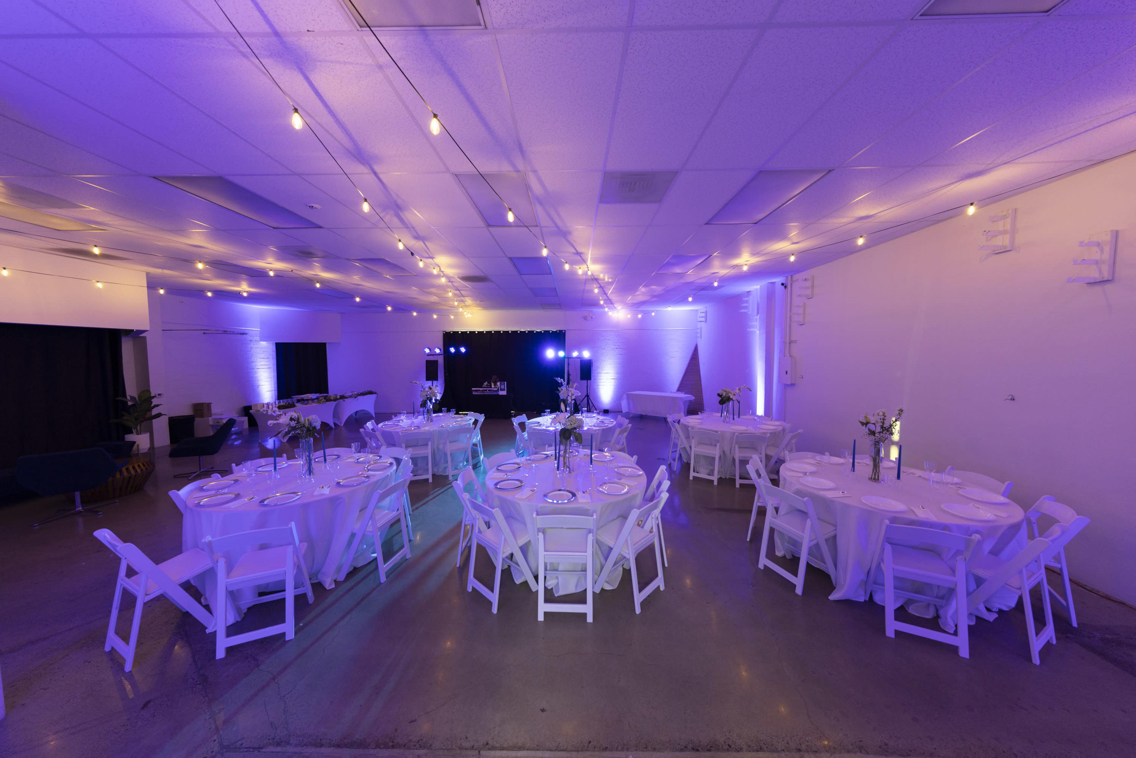 The image shows a spacious event venue set up with round tables and white chairs, illuminated by purple lighting and string lights.